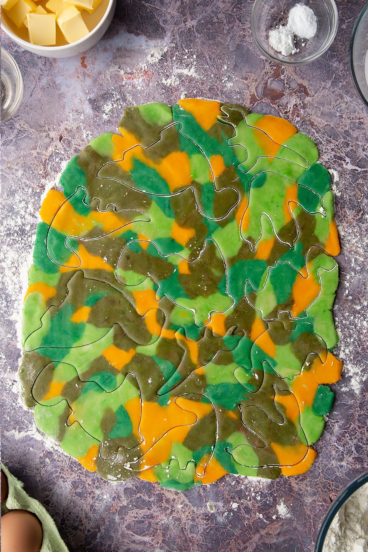 Overhead shot of small chunks of coloured cookie dough pieces combined and rolled out flat with dinosaur cookie shaped cut out