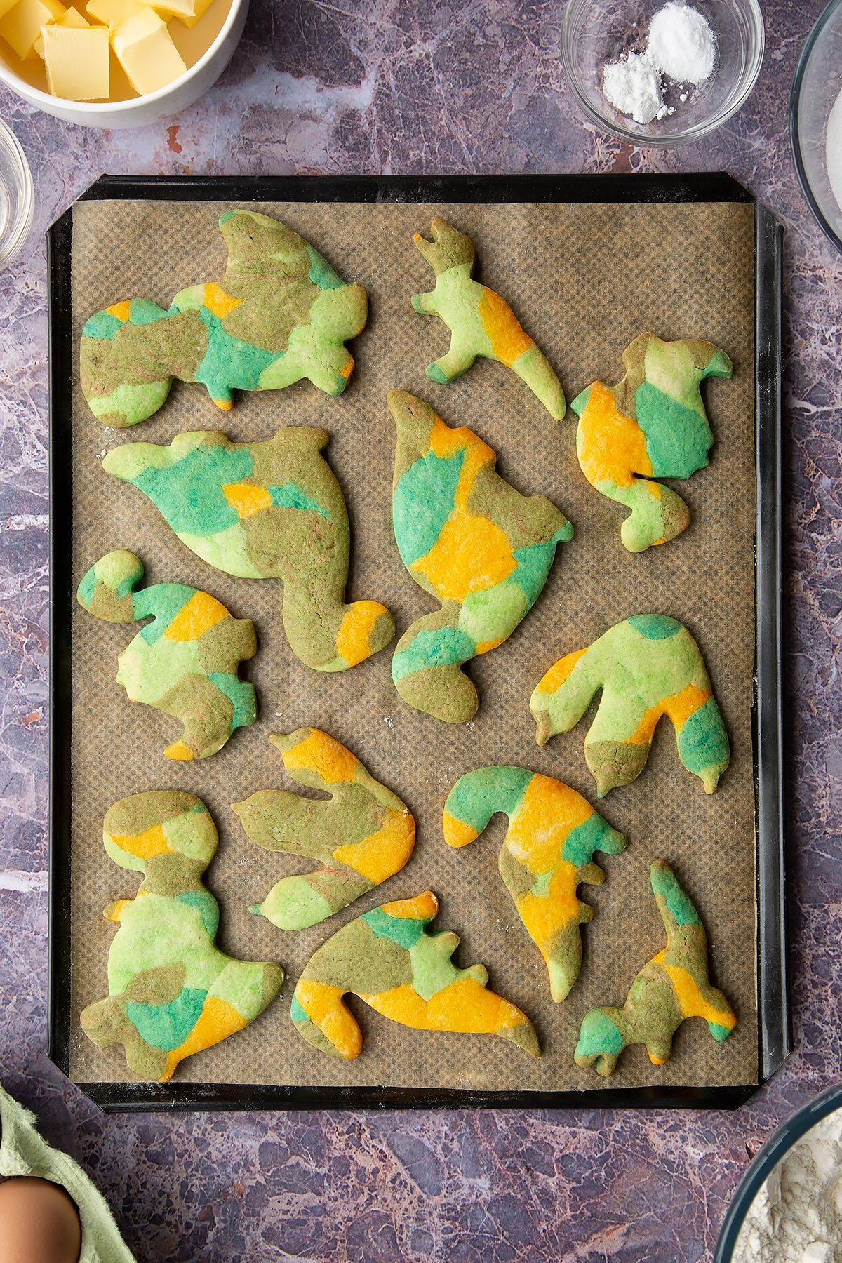 Overhead shot of baked multicoloured dinosaur cookie shapes on a lined baking tray