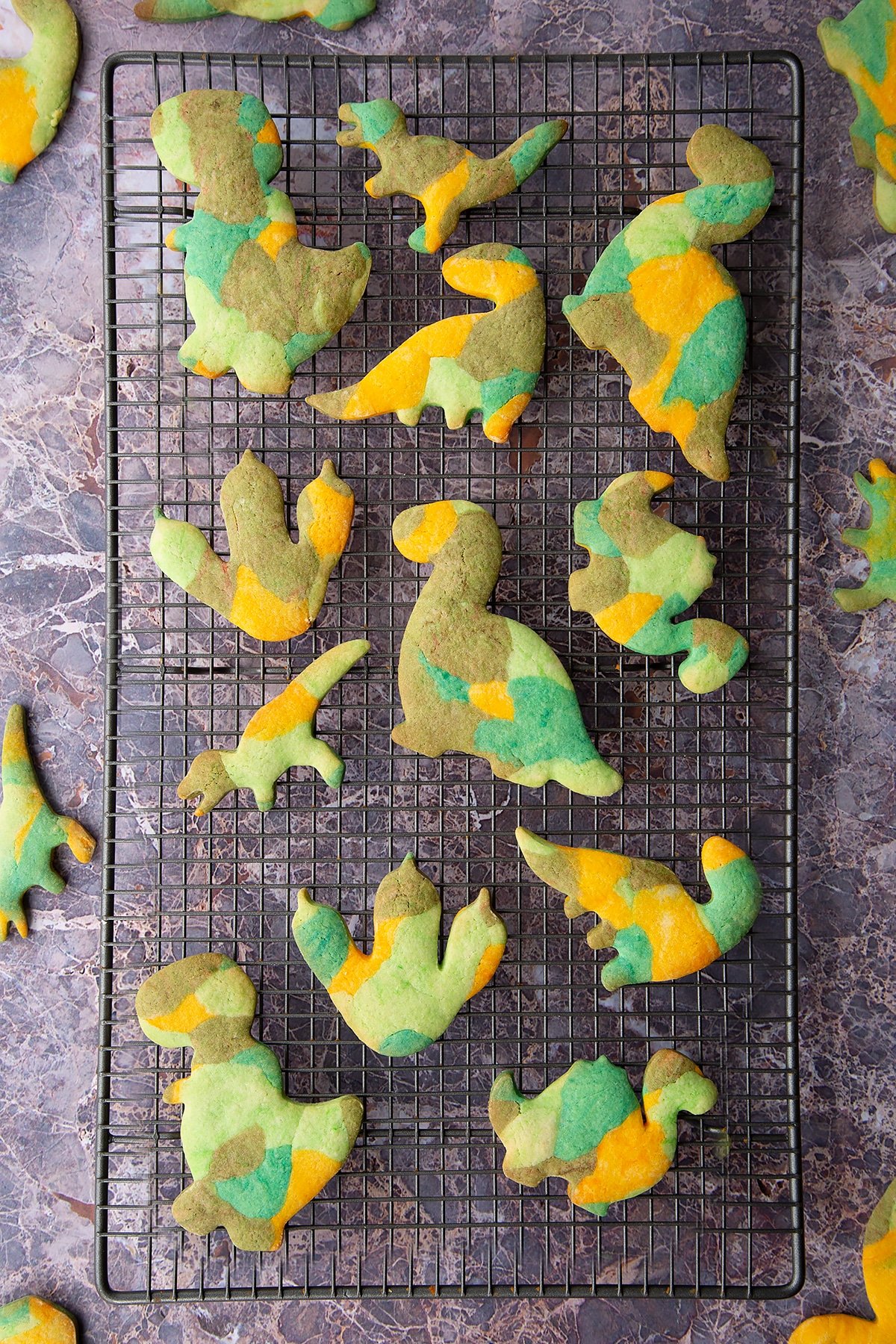 Overhead shot of baked multicoloured dinosaur cookie dough shapes on a metal rack