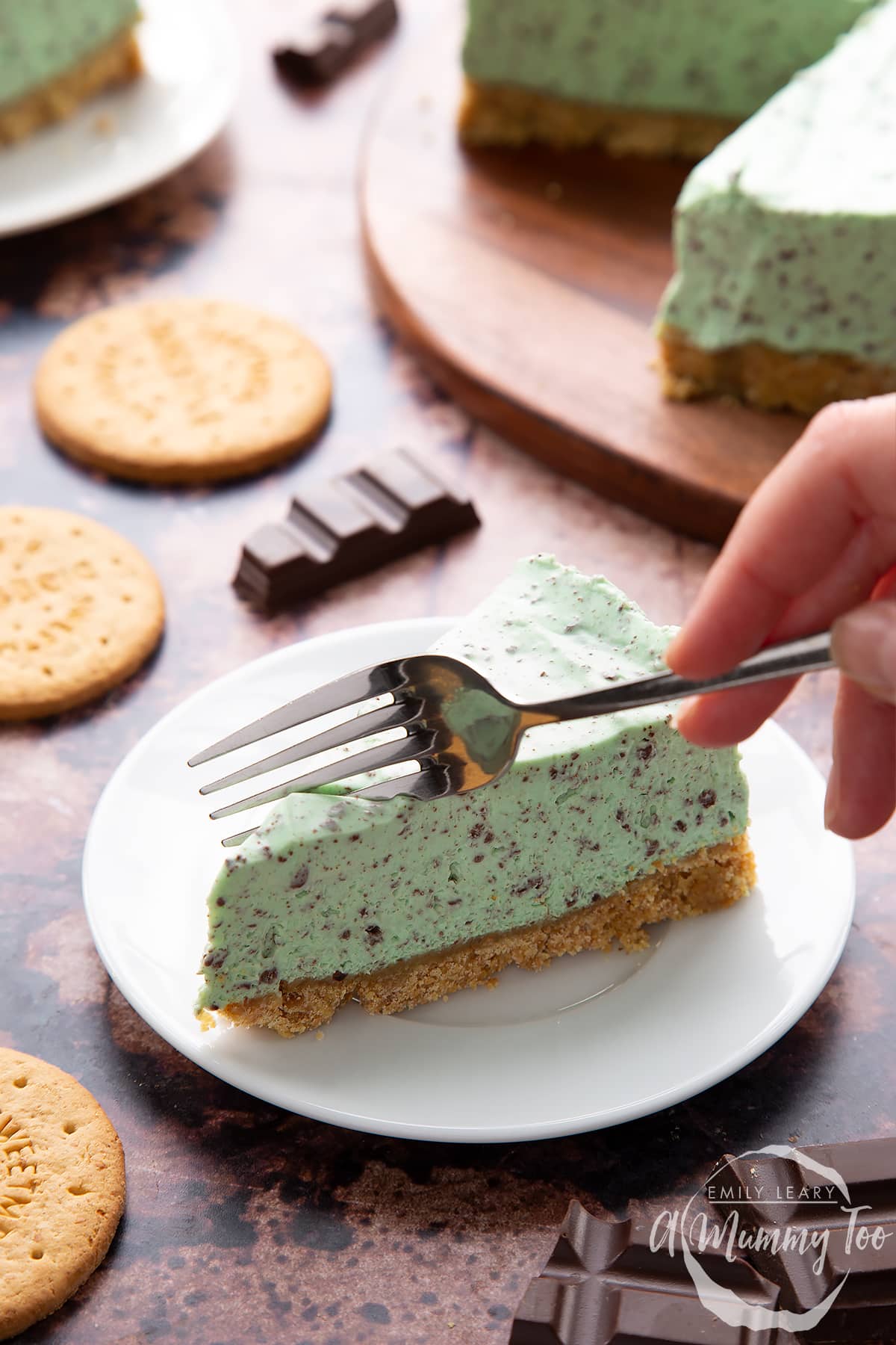 A slice of no bake mint cheesecake with a biscuit base on a white plate with a hand holding a fork.