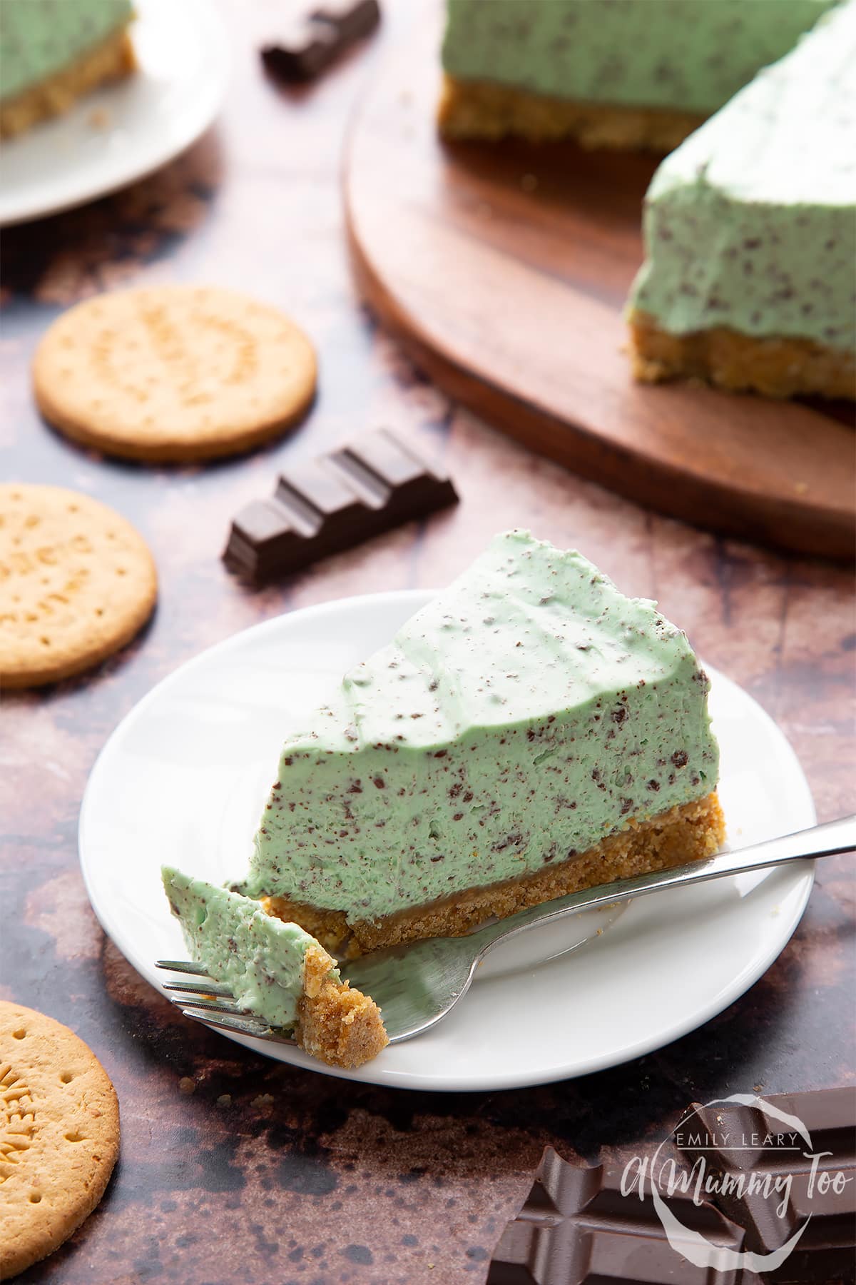A slice of no bake mint cheesecake with a biscuit base on a white plate. A fork rests on the plate with a piece of the cheesecake on it.