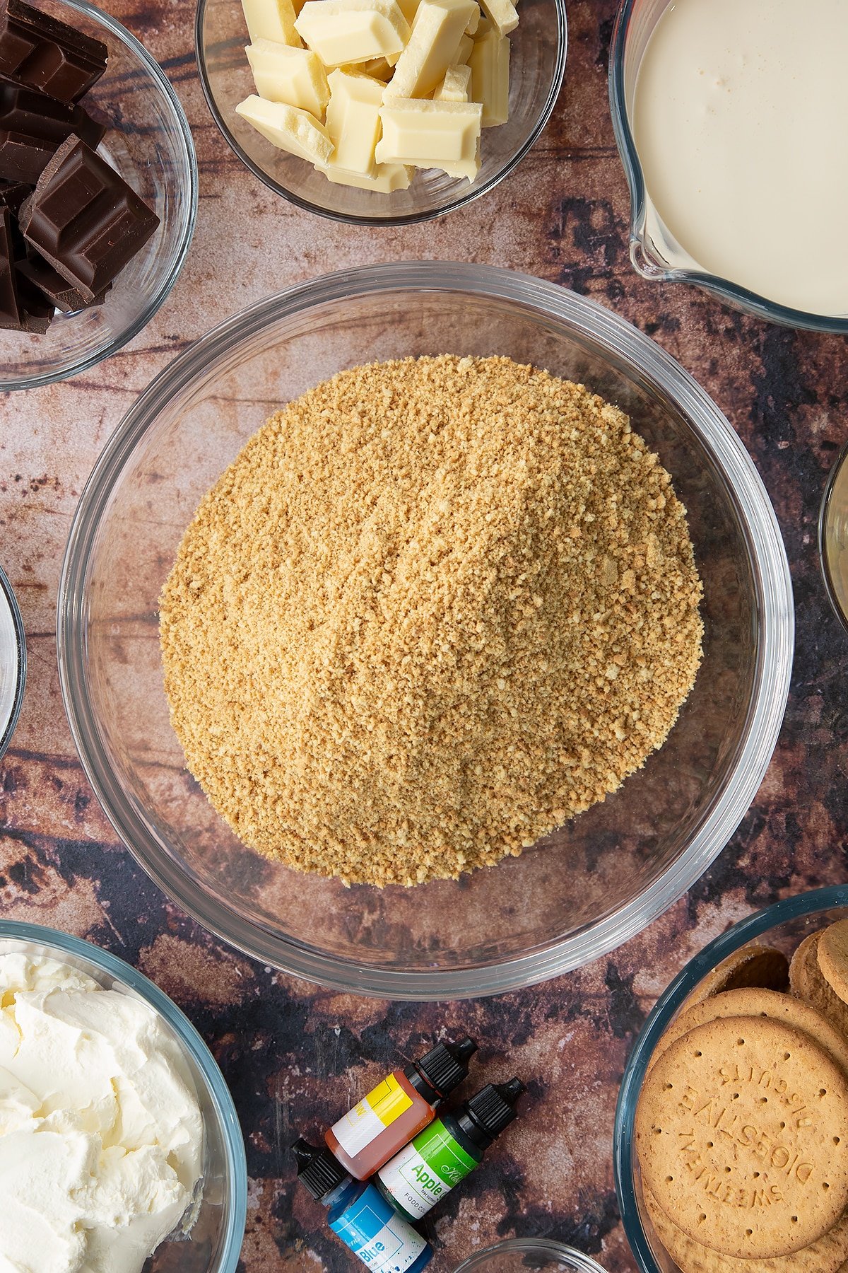 Crushed biscuits in a glass bowl. Ingredients to make no bake mint cheesecake surround the bowl.