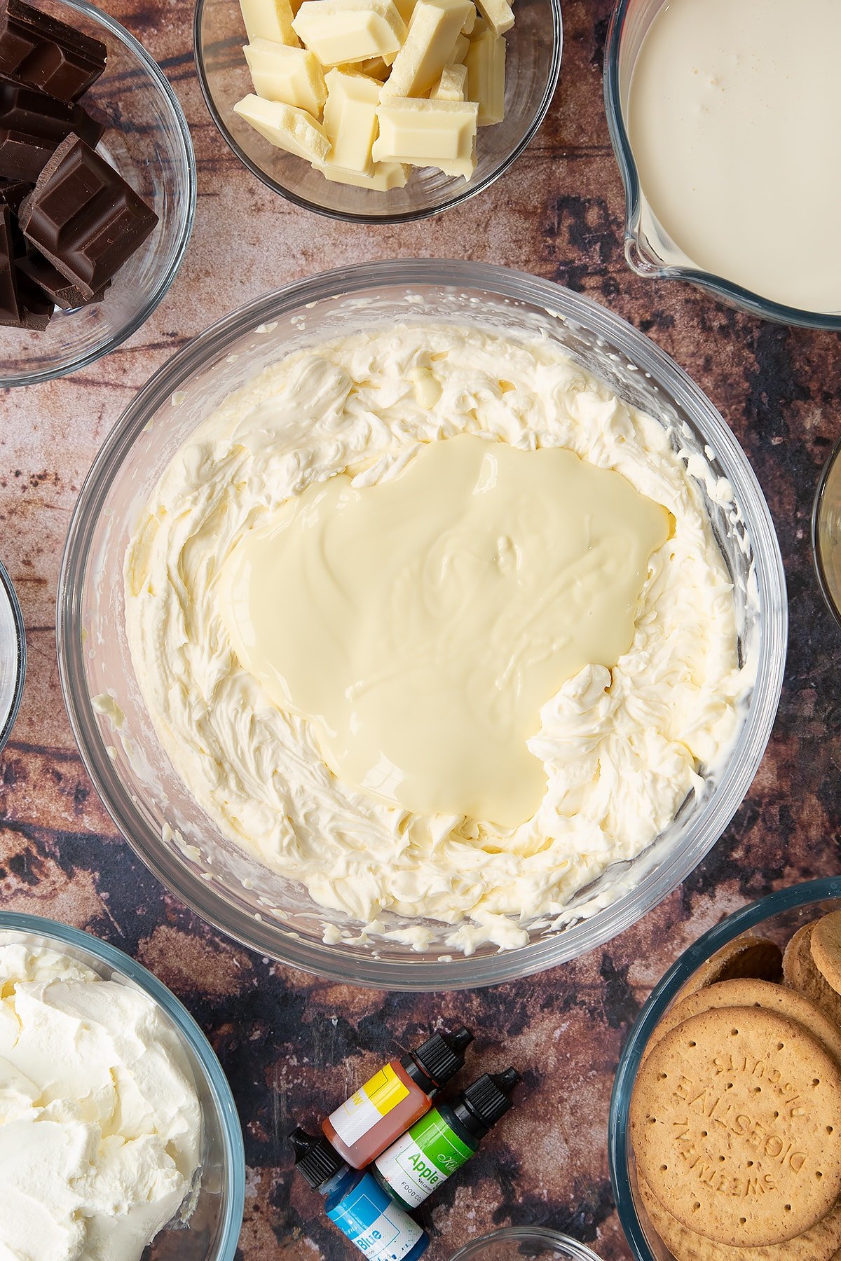 Double cream, cream cheese, peppermint extract and sugar, whisked together in a glass bowl with melted white chocolate on top. Ingredients to make no bake mint cheesecake surround the bowl.