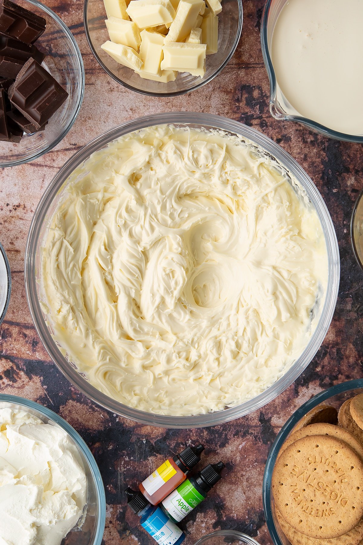 Cheesecake filling in a glass bowl. Ingredients to make no bake mint cheesecake surround the bowl.