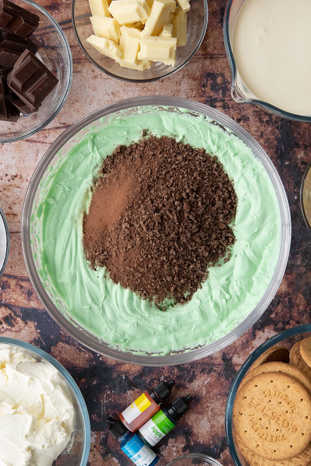 Peppermint green cheesecake filling in a glass bowl with chopped dark chocolate on top. Ingredients to make no bake mint cheesecake surround the bowl.