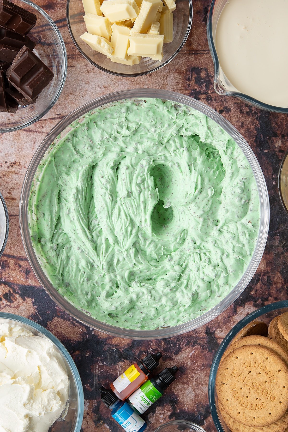 Peppermint green, chocolate chip cheesecake filling in a glass bowl. Ingredients to make no bake mint cheesecake surround the bowl.