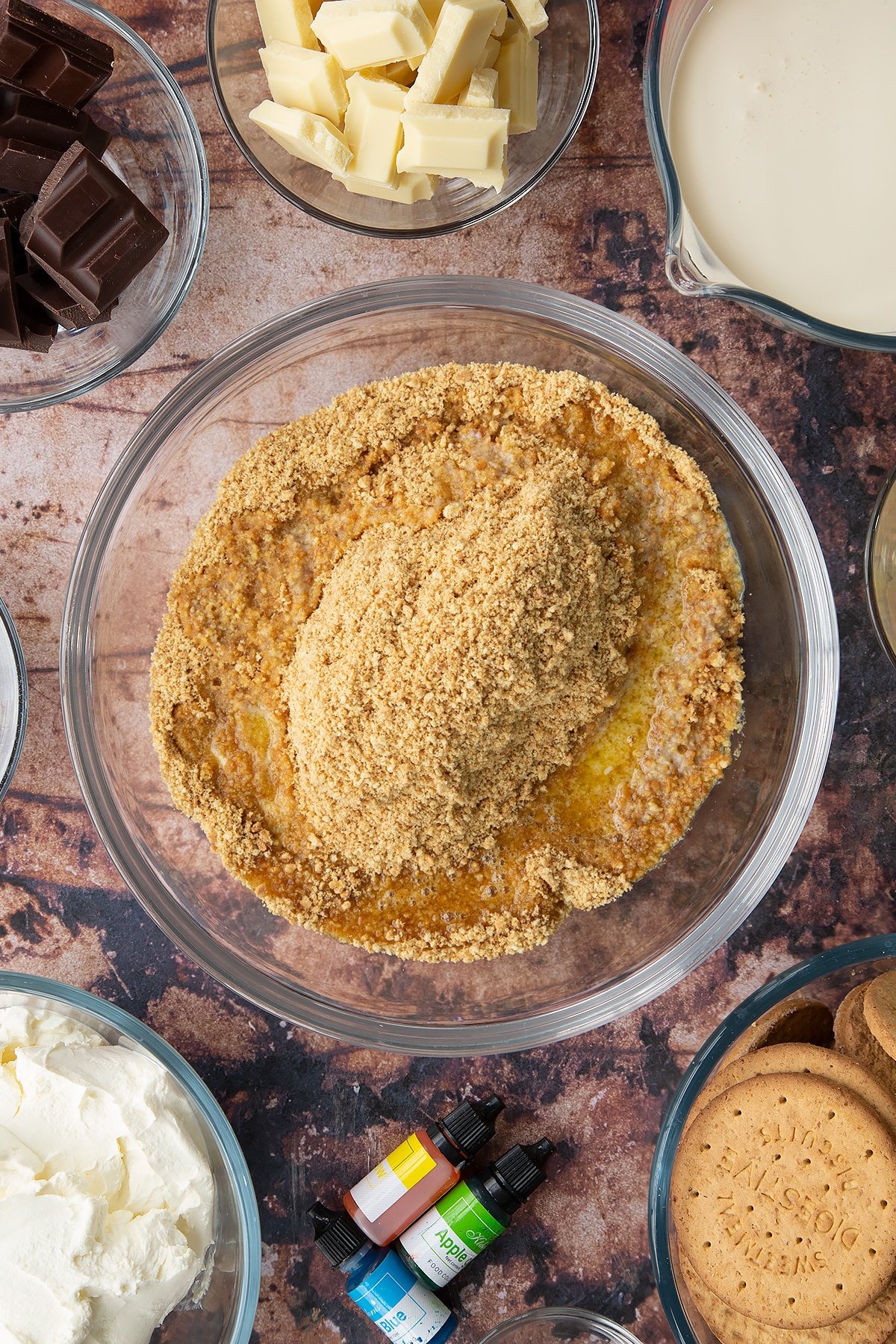 Crushed biscuits and melted butter in a glass bowl. Ingredients to make no bake mint cheesecake surround the bowl.