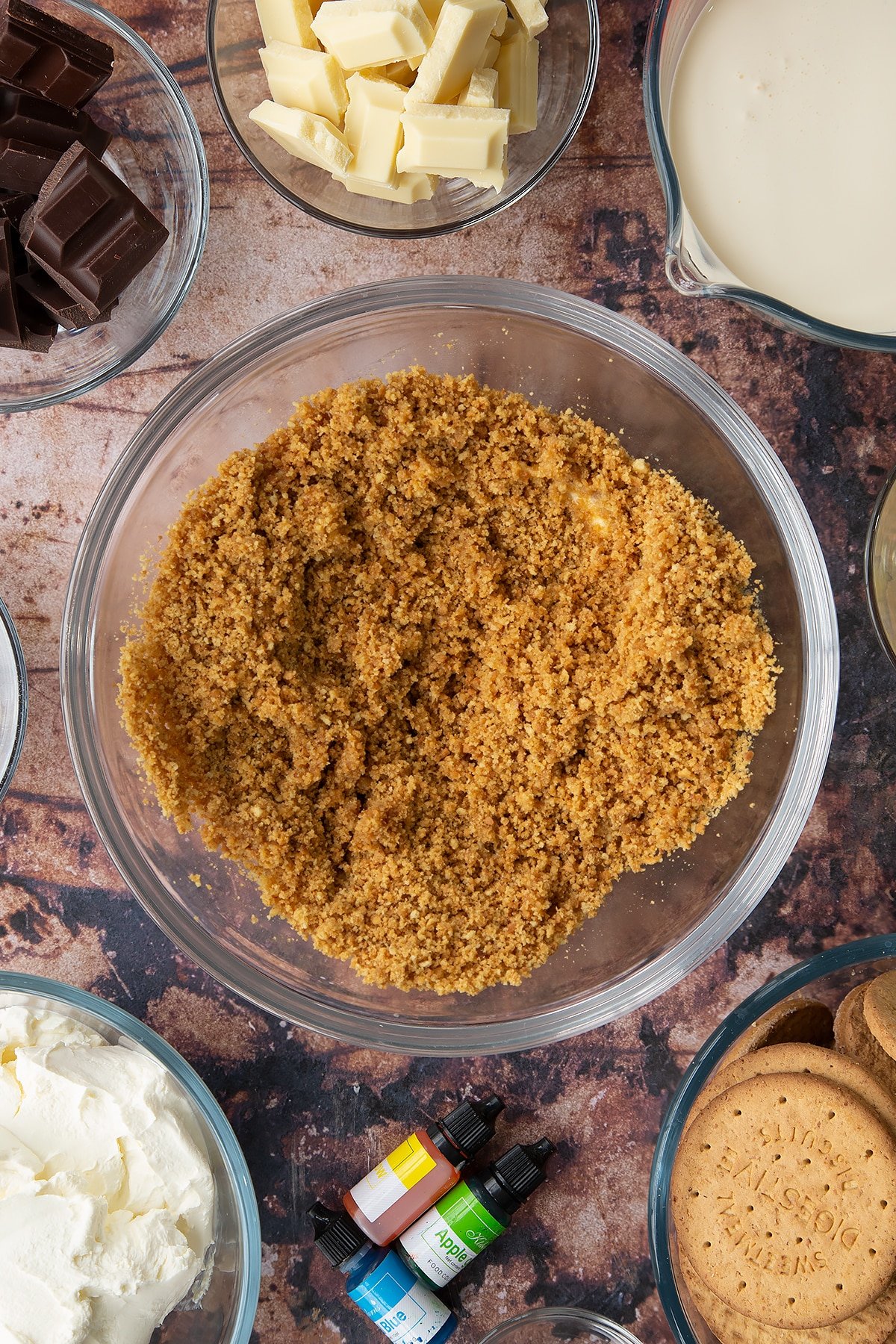 Crushed biscuits mixed with melted butter in a glass bowl. Ingredients to make no bake mint cheesecake surround the bowl.