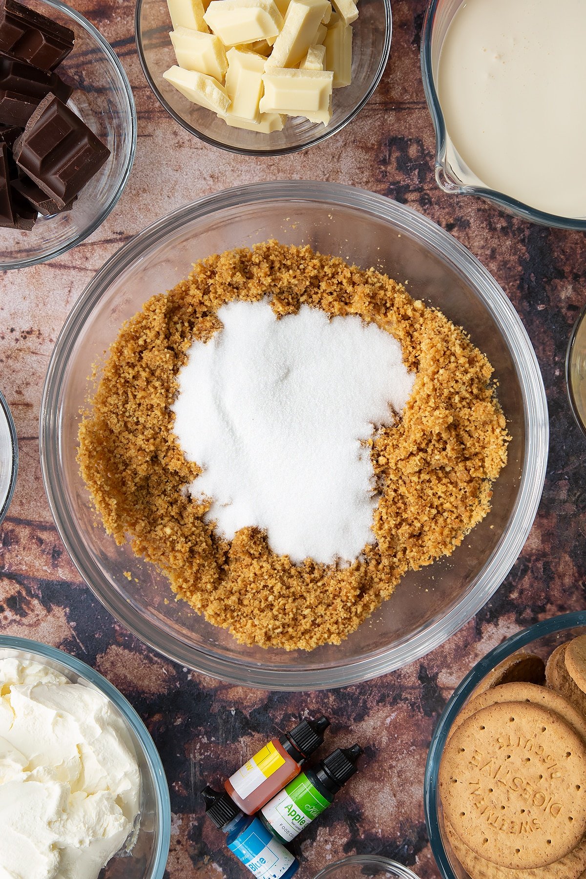 Crushed biscuits mixed with melted butter in a glass bowl with sugar on top. Ingredients to make no bake mint cheesecake surround the bowl.