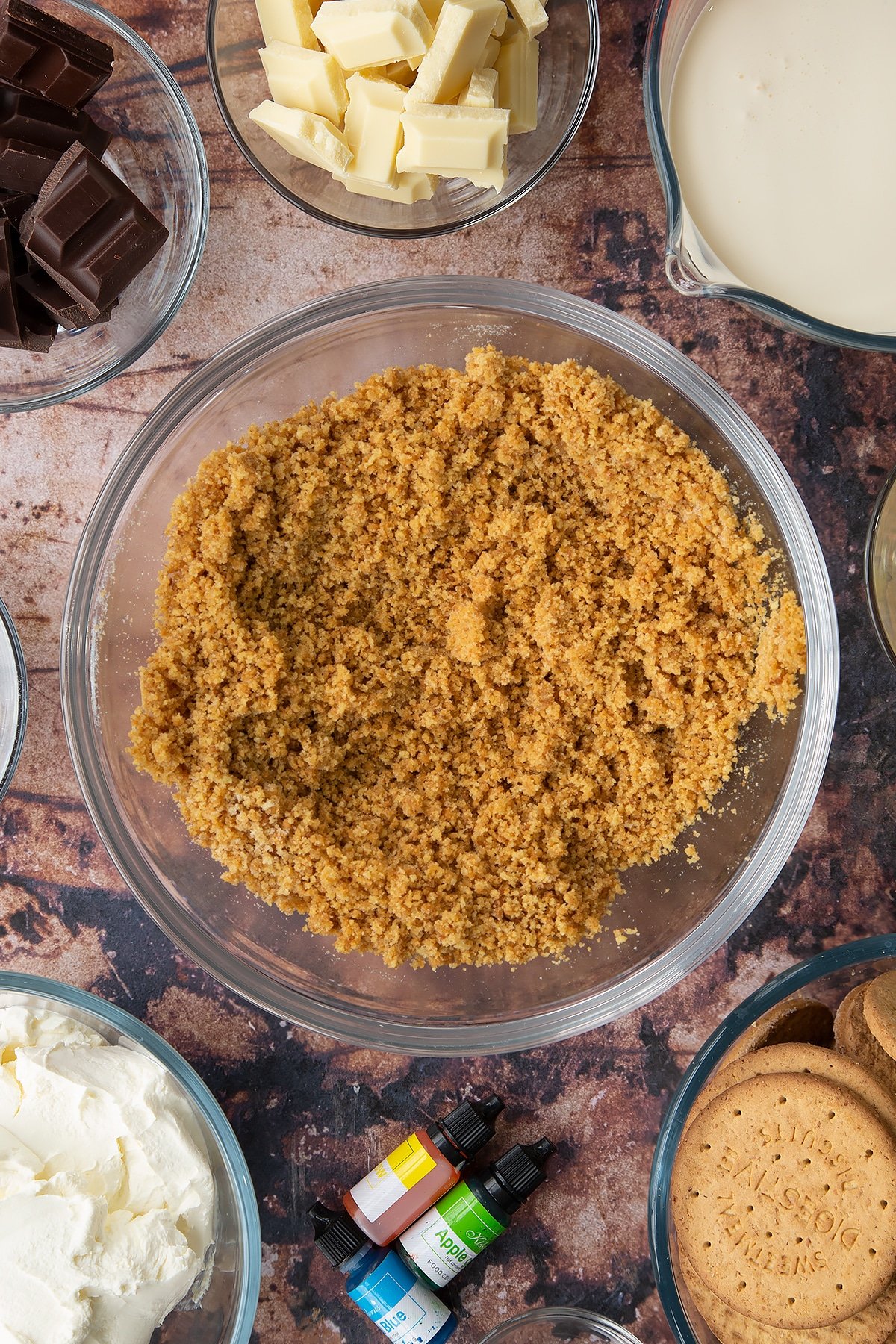 Crushed biscuits mixed with sugar and melted butter in a glass bowl. Ingredients to make no bake mint cheesecake surround the bowl.