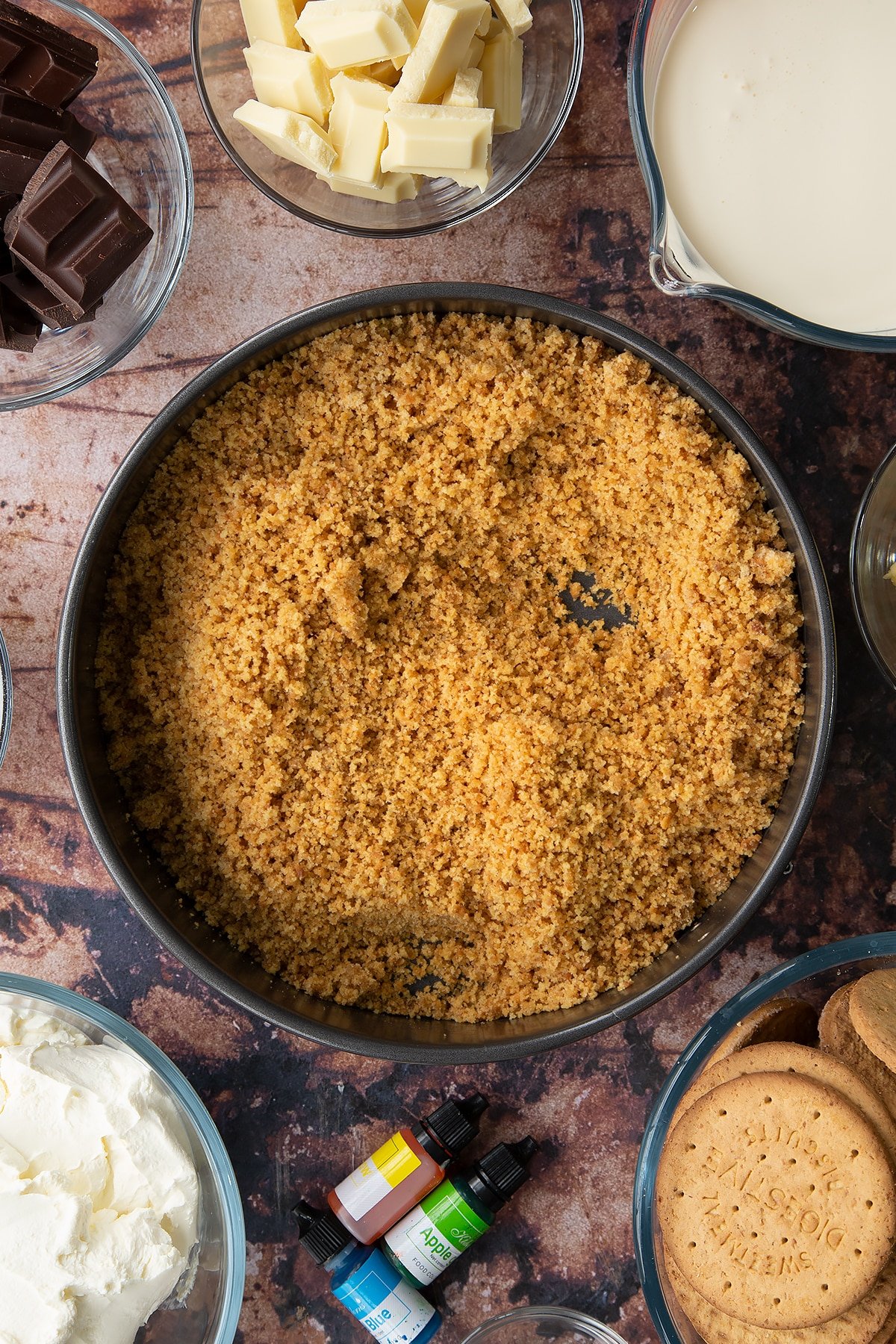 Crushed biscuits mixed with sugar and melted butter in a baking tin. Ingredients to make no bake mint cheesecake surround the bowl.