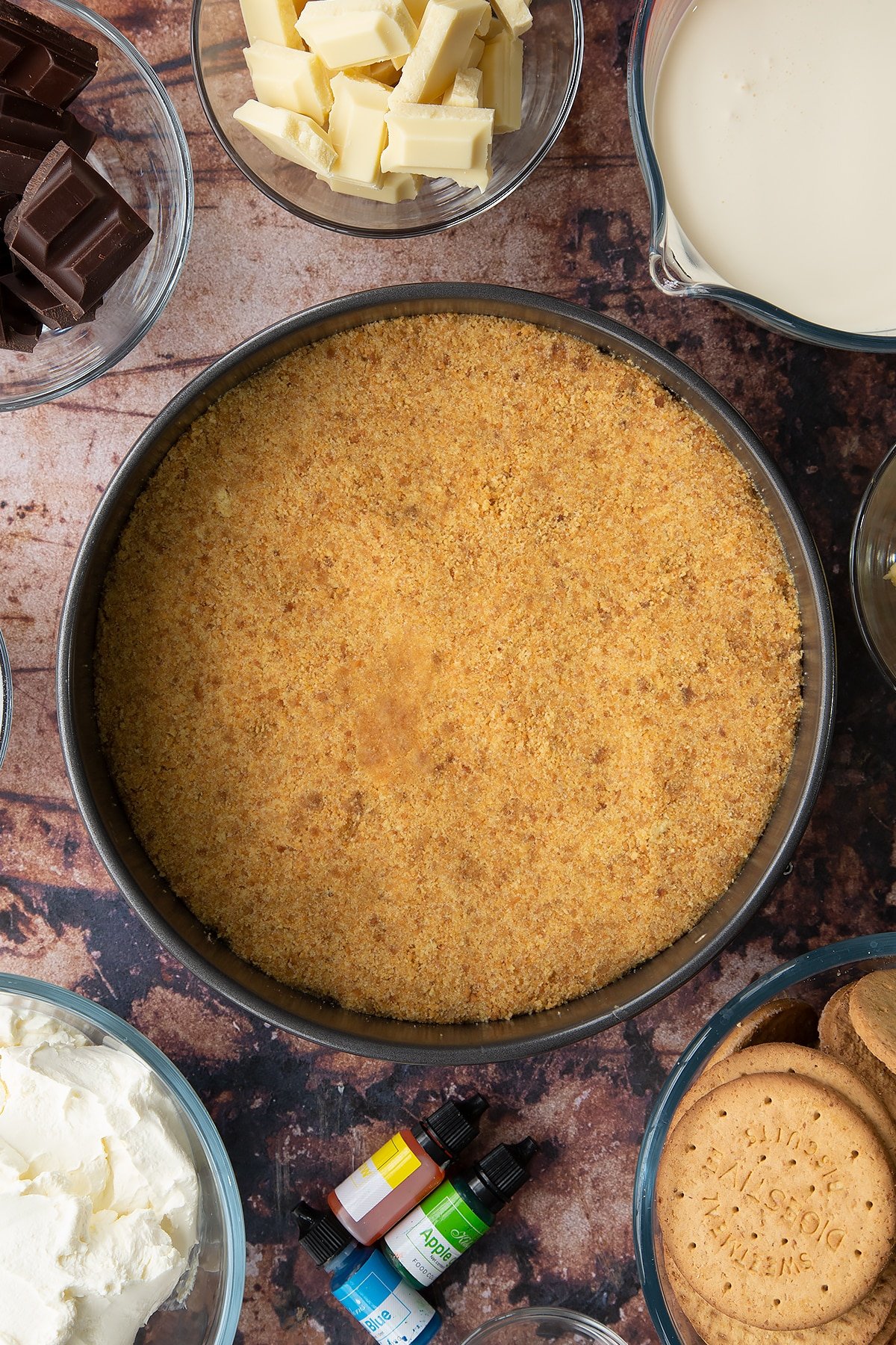 Crushed biscuits mixed with sugar and melted butter pressed into the base of a baking tin. Ingredients to make no bake mint cheesecake surround the bowl.