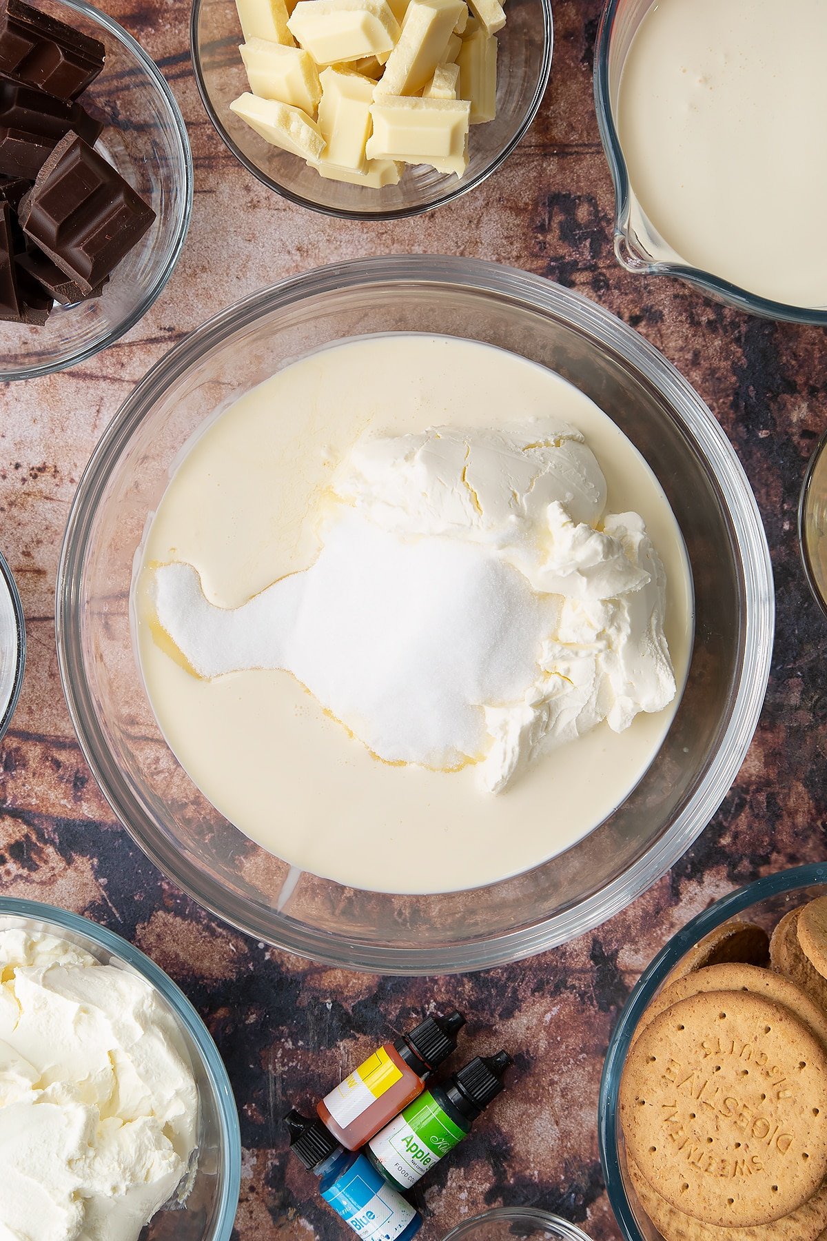 Double cream, cream cheese, peppermint extract and sugar in a glass bowl. Ingredients to make no bake mint cheesecake surround the bowl.