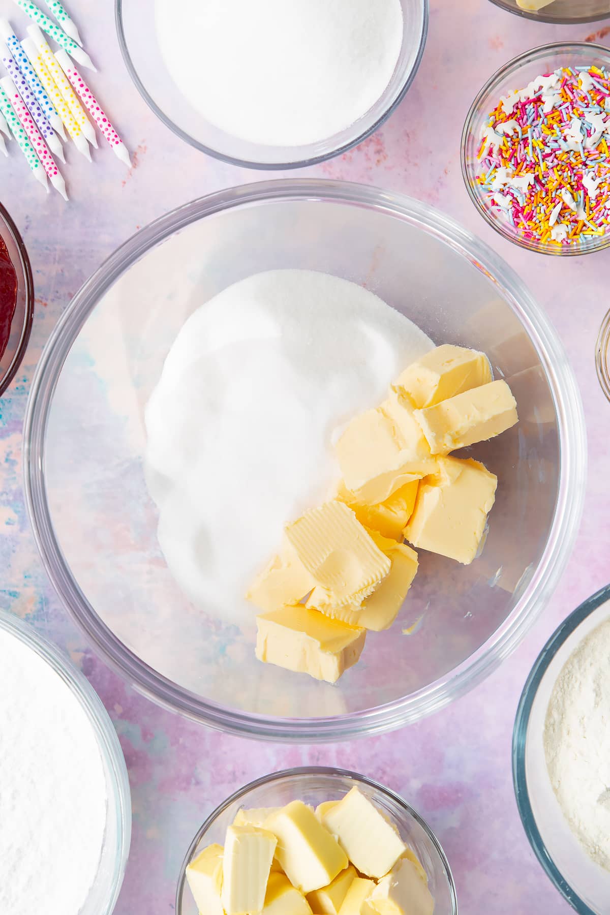 Vegan butter and caster sugar in a glass mixing bowl. Ingredients to make vegan birthday cake surround the bowl.