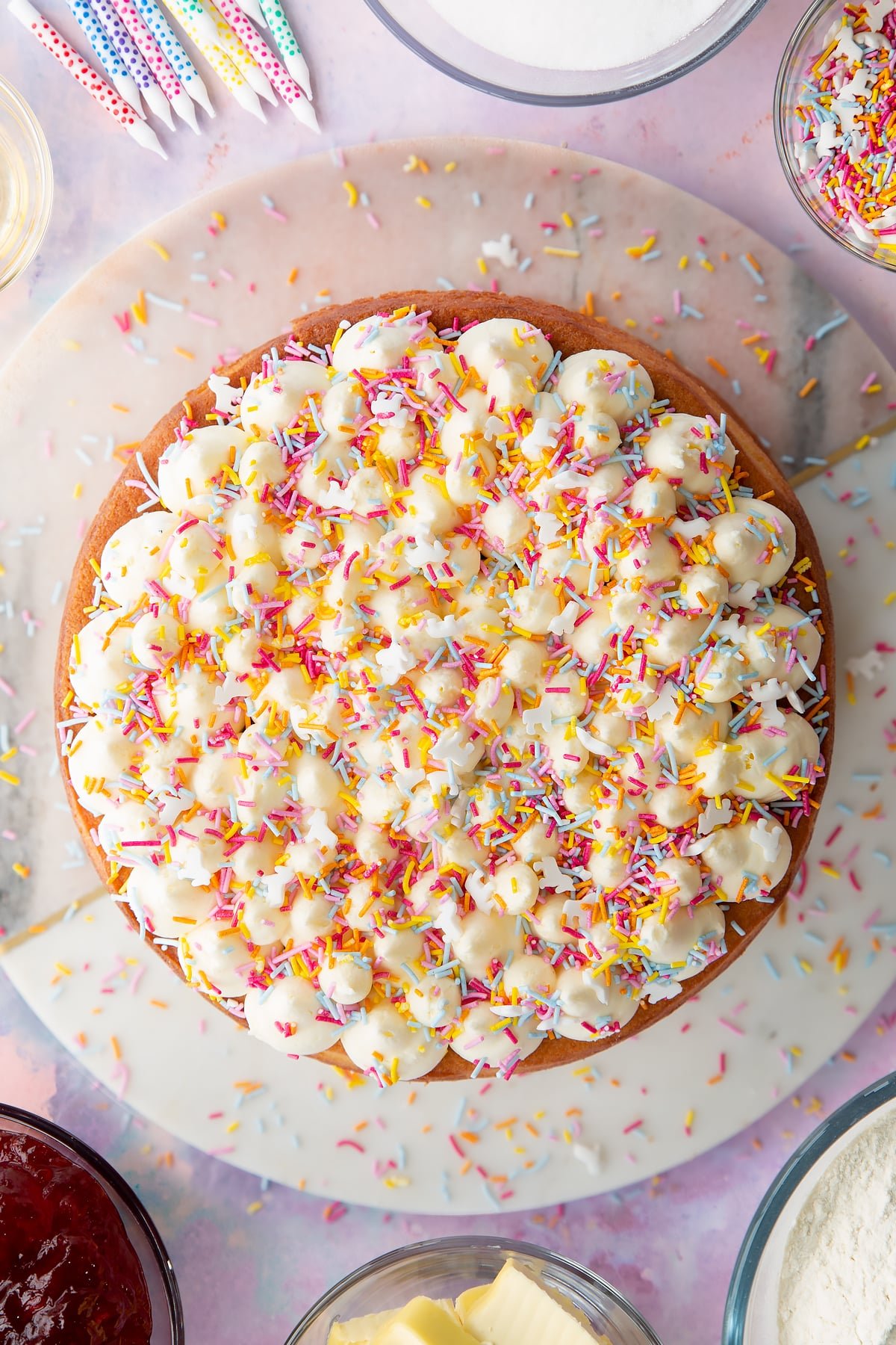 Three vegan vanilla sponges, sandwiched with buttercream and jam and topped with buttercream and sprinkles on a pink and white round marble board.