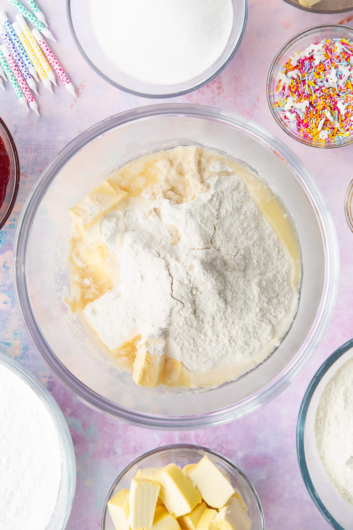 Vegan butter, caster sugar, self-raising flour, vanilla, plant milk and aquafaba in a glass mixing bowl. Ingredients to make vegan birthday cake surround the bowl.