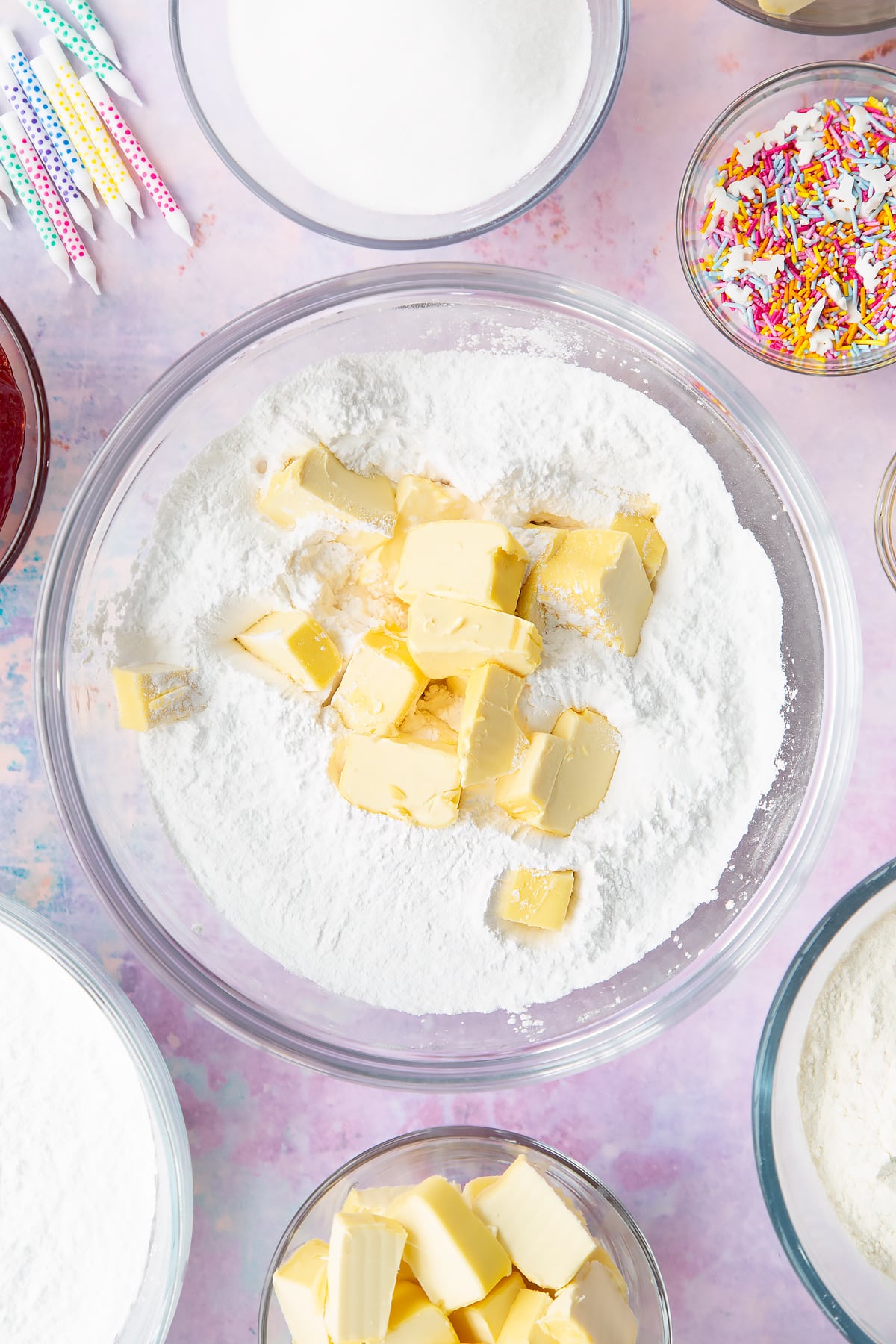 Vegan butter, icing sugar, vanilla extract and plant milk in a glass mixing bowl. Ingredients to make vegan birthday cake surround the bowl.