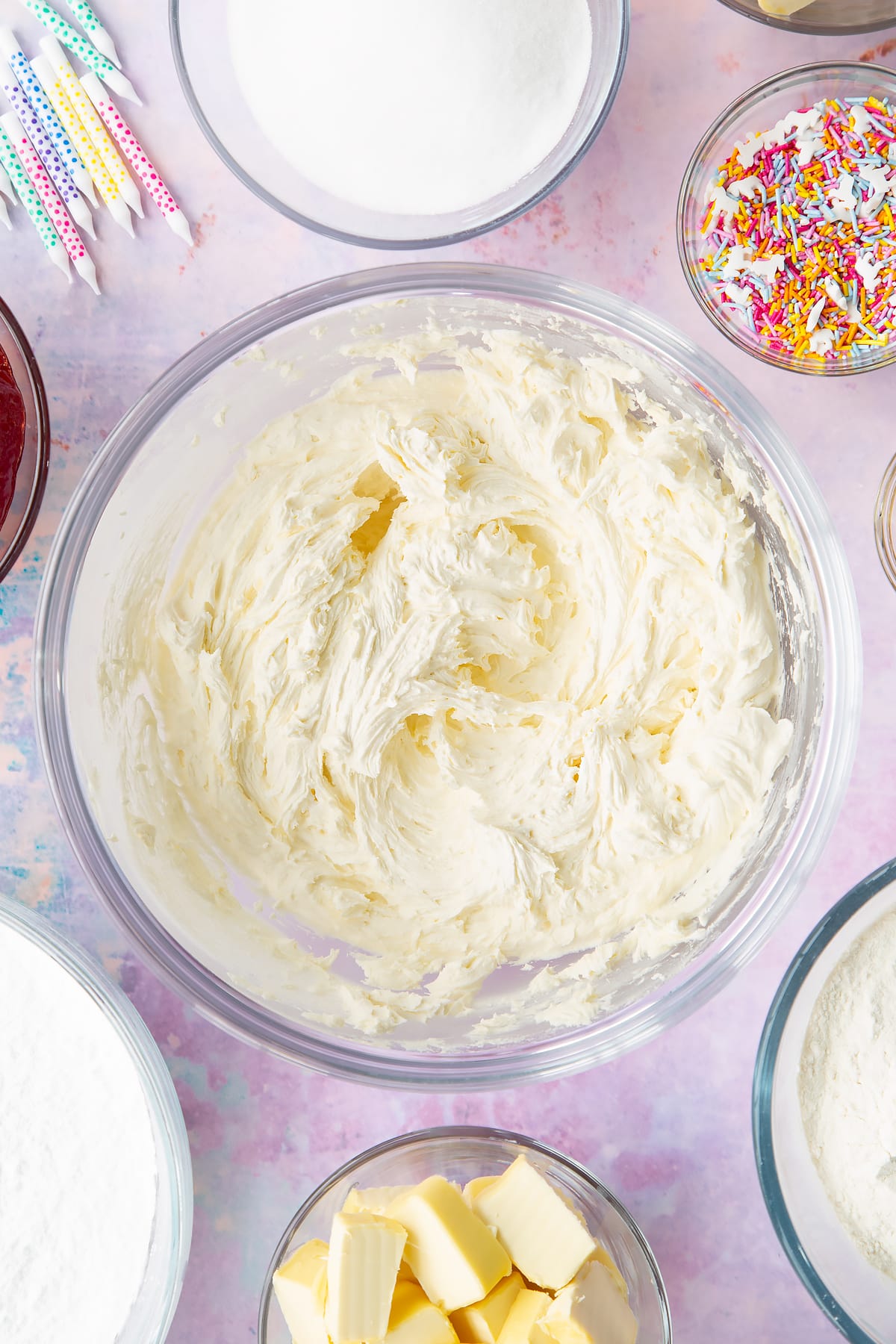 Vegan buttercream frosting in a glass mixing bowl. Ingredients to make vegan birthday cake surround the bowl.