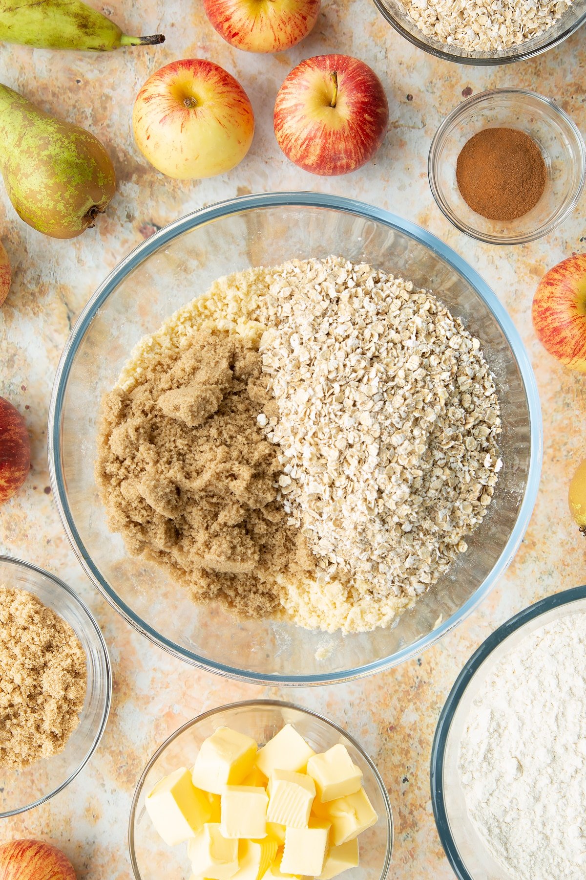 Flour and butter rubbed together in a mixing bowl with sugar and oats on top. Ingredients to make easy apple pear crumble surround the bowl.