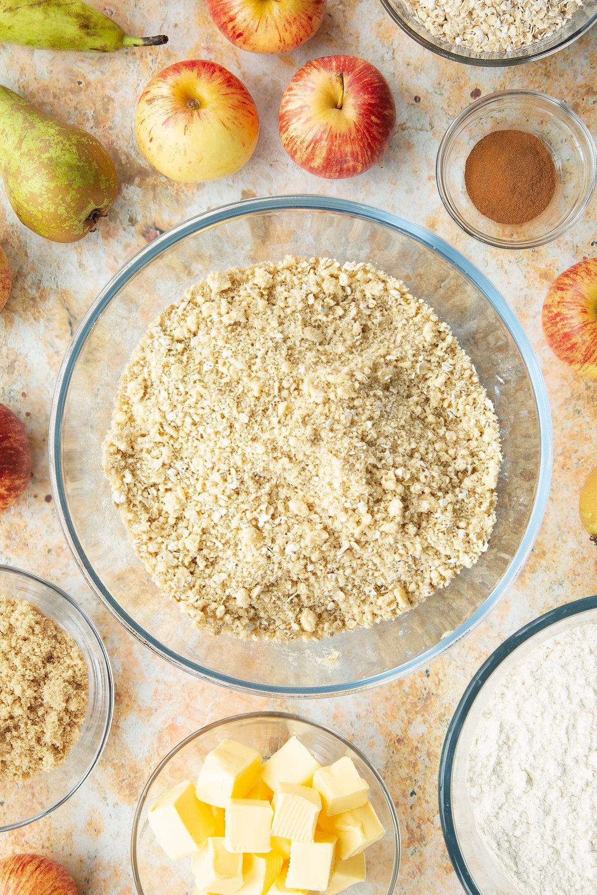 Flour, butter oats and sugar mixed together in a mixing bowl. Ingredients to make easy apple pear crumble surround the bowl.