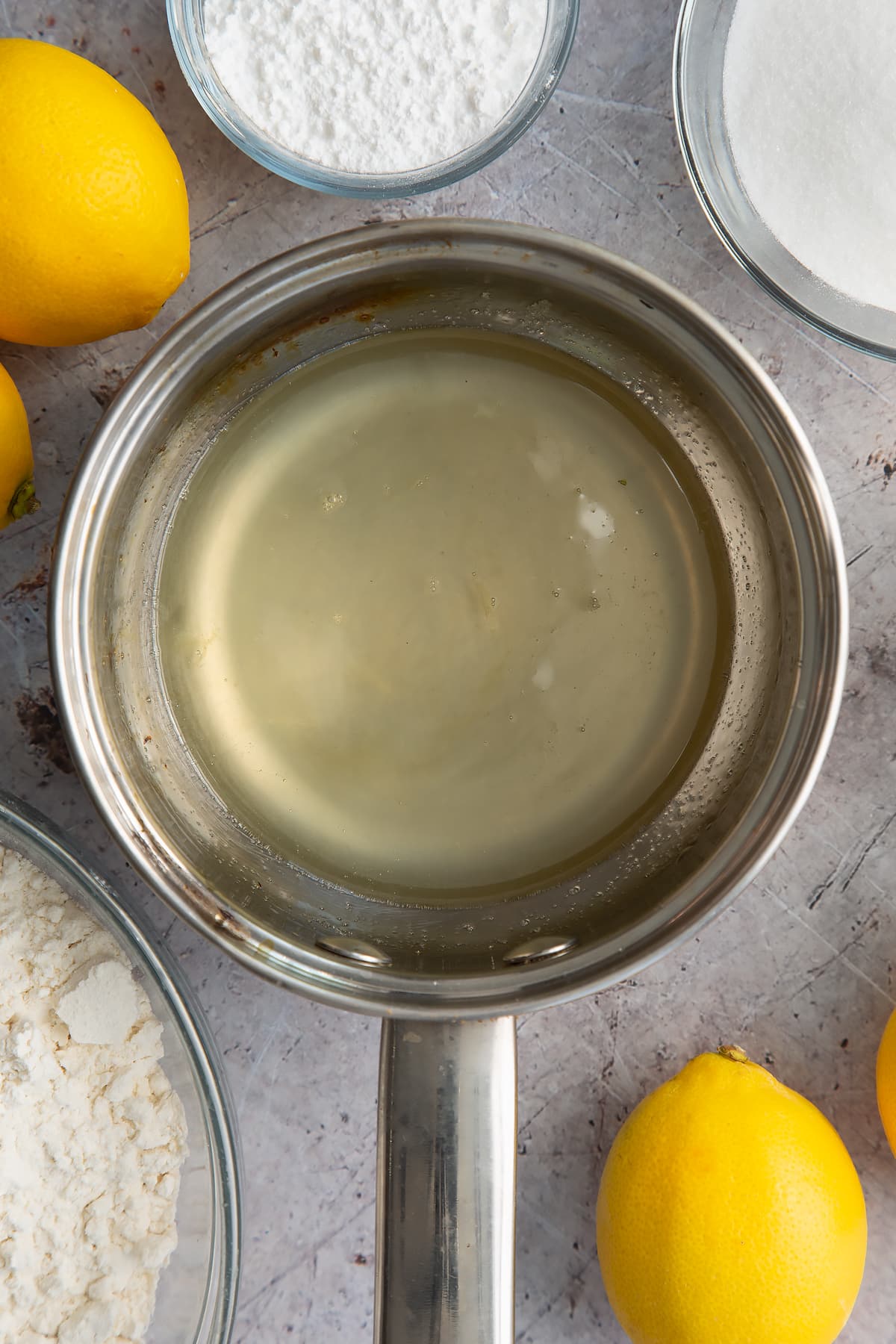 Hot lemon drizzle syrup in a small metal pan. Ingredients to make lemon drizzle cake surround the pan.