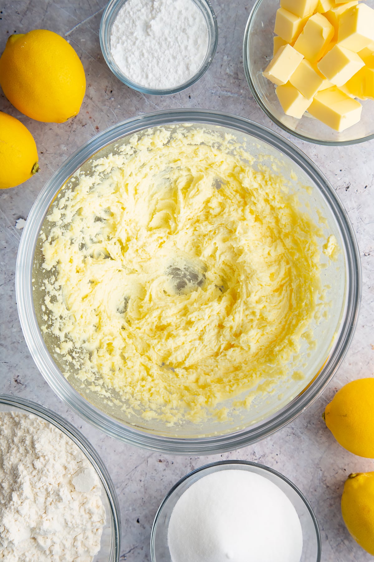 Butter and sugar creamed together in a glass bowl. Ingredients to make lemon drizzle cake surround the bowl. 
