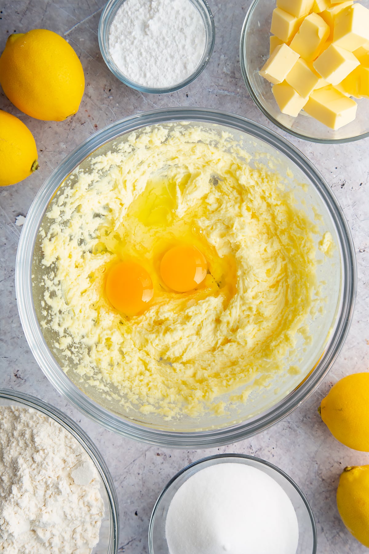 Butter and sugar creamed together with eggs on top in a glass bowl. Ingredients to make lemon drizzle cake surround the bowl.