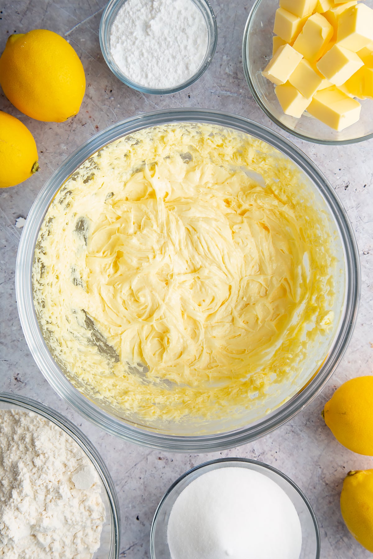 Butter, sugar and eggs creamed together in a glass bowl. Ingredients to make lemon drizzle cake surround the bowl. 