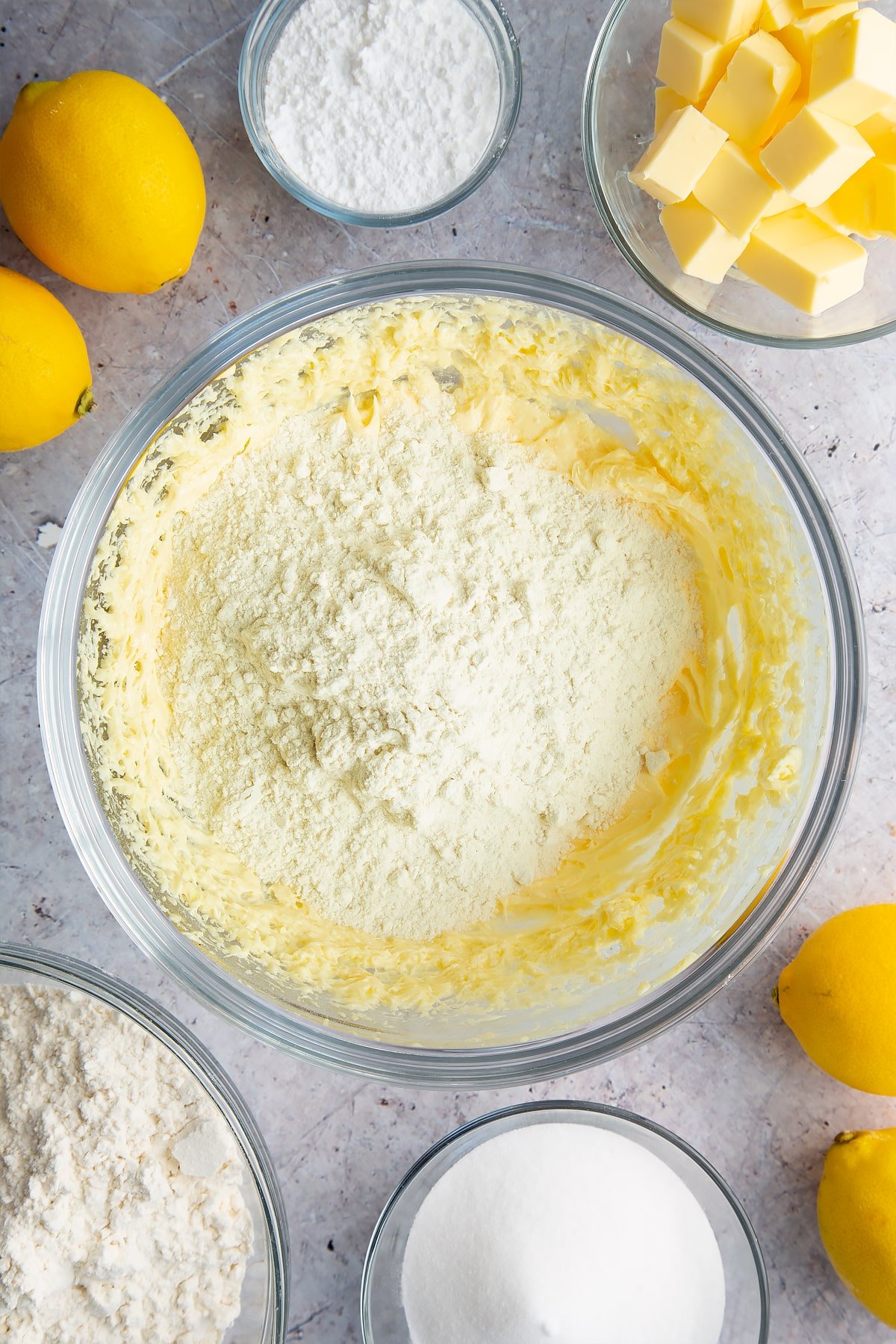 Butter, sugar and eggs creamed together with flour on top in a glass bowl. Ingredients to make lemon drizzle cake surround the bowl. 