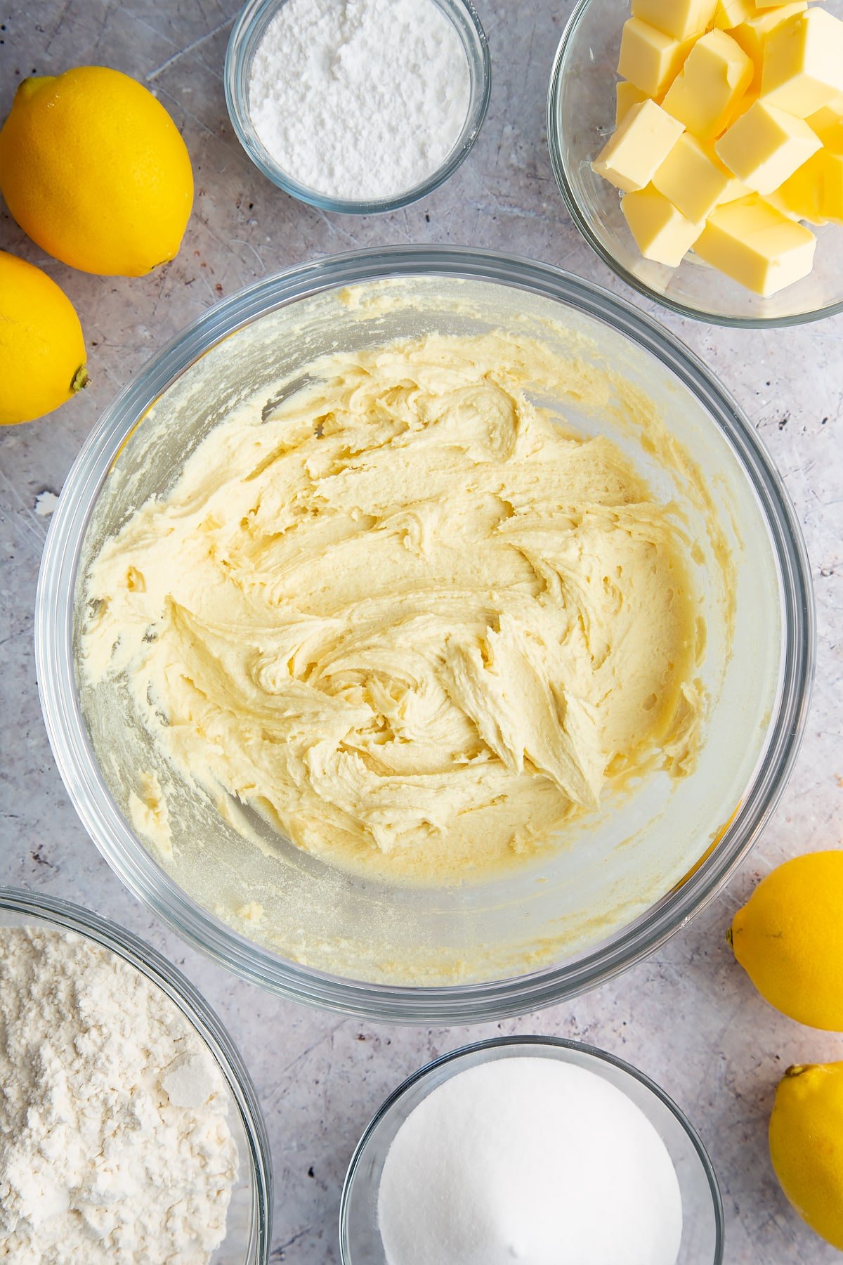 Cake batter in a glass bowl. Ingredients to make lemon drizzle cake surround the bowl. 