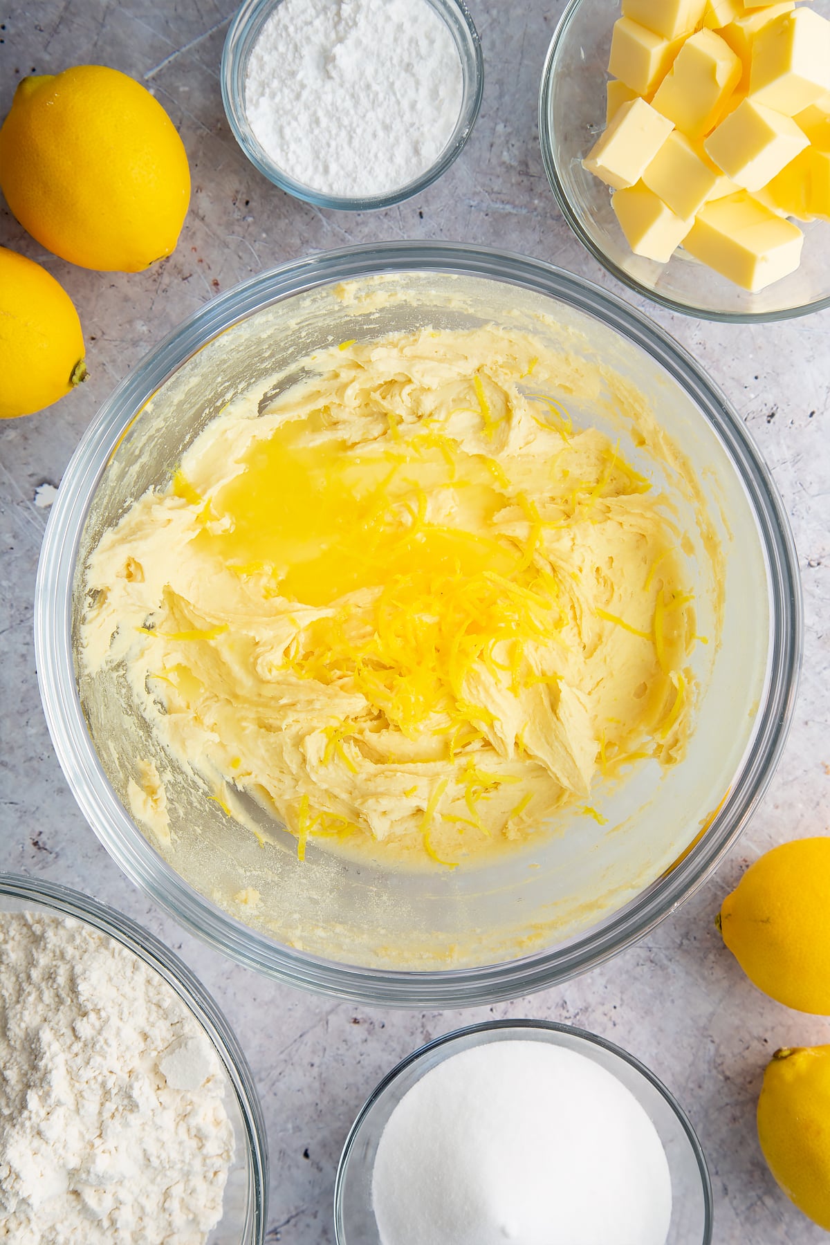 Cake batter with lemon juice and zest on top in a glass bowl. Ingredients to make lemon drizzle cake surround the bowl. 