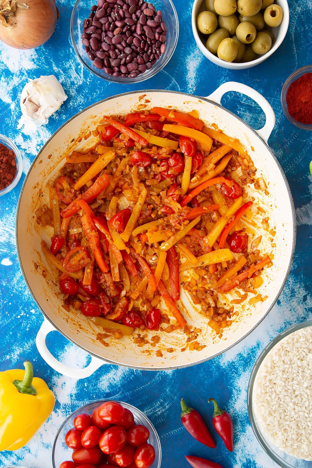 Sweated onions, spices, tomatoes and mixed sliced peppers in a pan. Ingredients to make easy Spanish rice and beans surround the pan.