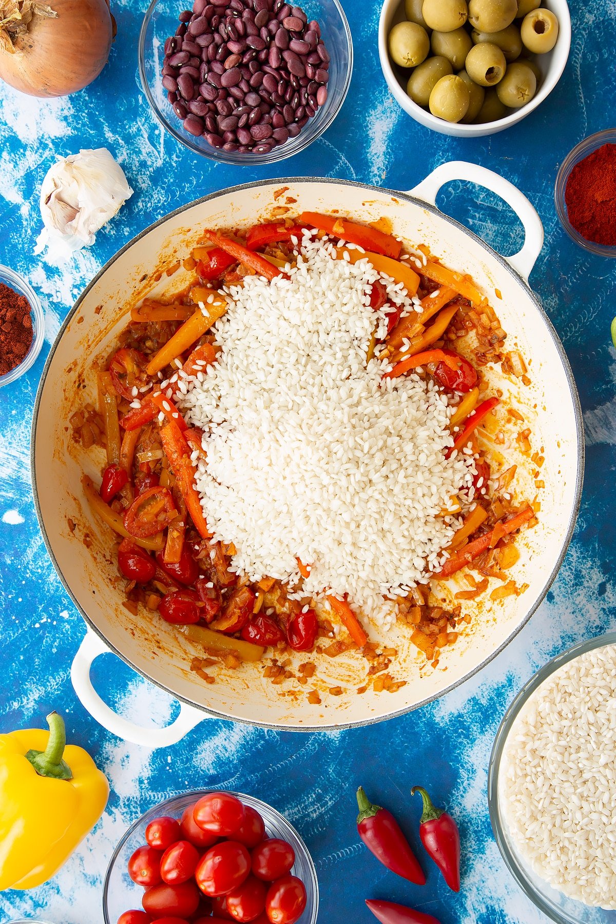 Sweated onions, spices, tomatoes and mixed sliced peppers in a pan with arborio rice on top. Ingredients to make easy Spanish rice and beans surround the pan.
