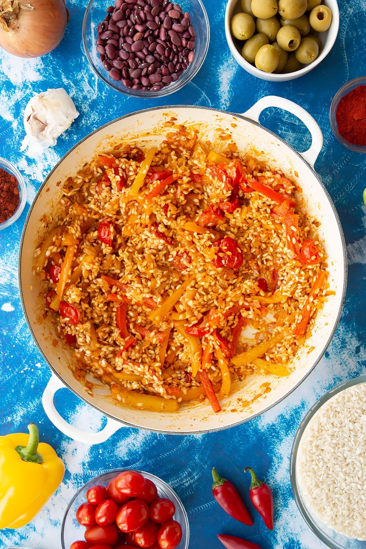 Sweated onions, spices, tomatoes and mixed sliced peppers in a pan with arborio rice mixed in. Ingredients to make easy Spanish rice and beans surround the pan.