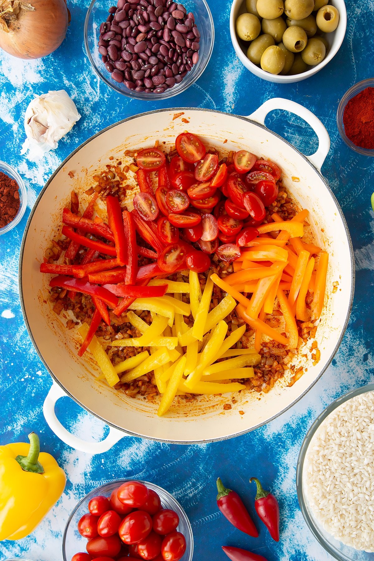 Sweated onions, garlic, cayenne, paprika and oregano in a pan with tomatoes and mixed sliced peppers. Ingredients to make easy Spanish rice and beans surround the pan.