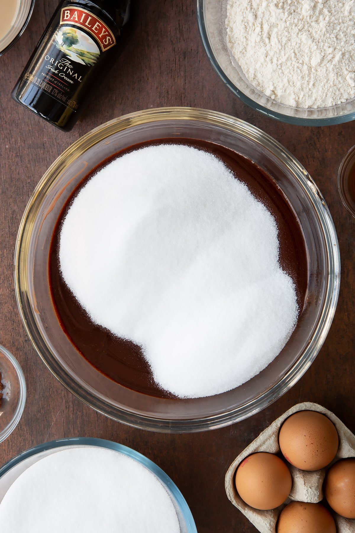 Melted chocolate and butter in a glass bowl with sugar on top. Ingredients to make Baileys brownies surround the bowl.