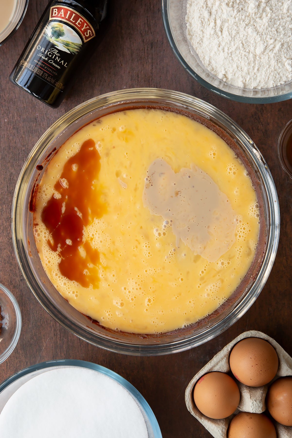 Melted chocolate, butter and sugar in a glass bowl with beaten eggs, vanilla and Baileys on top. Ingredients to make Baileys brownies surround the bowl.