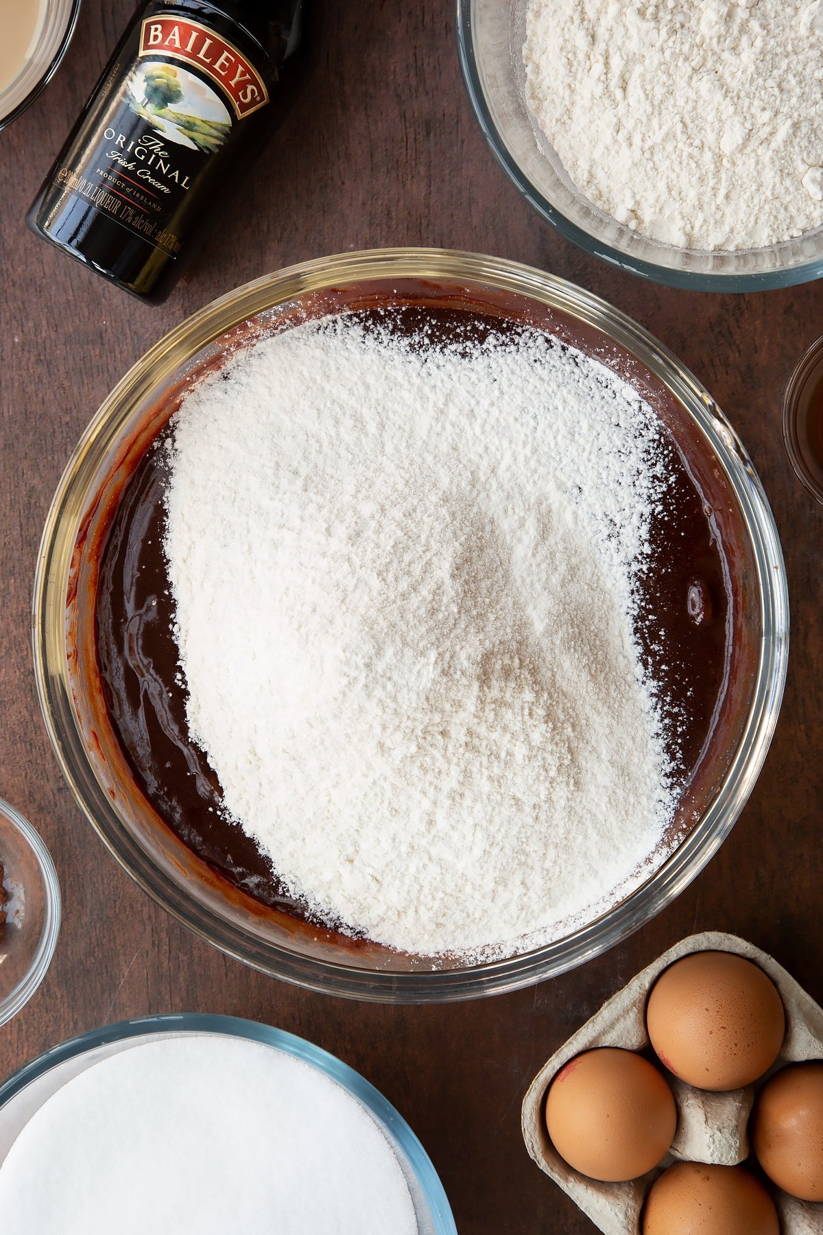 Melted chocolate, butter, Baileys, sugar and eggs in a glass bowl with flour on top. Ingredients to make Baileys brownies surround the bowl.