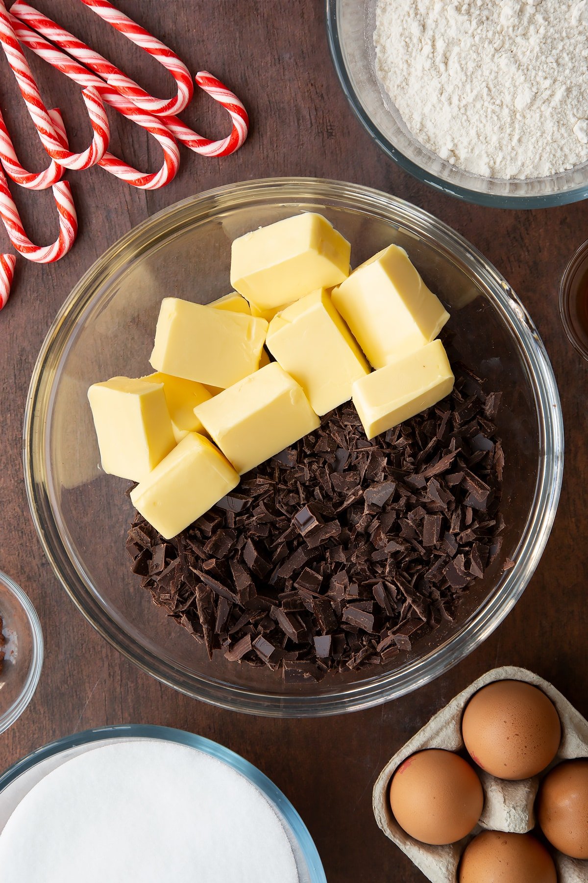 Cubed butter and chopped dark chocolate in a glass bowl. Ingredients to make Candy cane brownies surround the bowl.