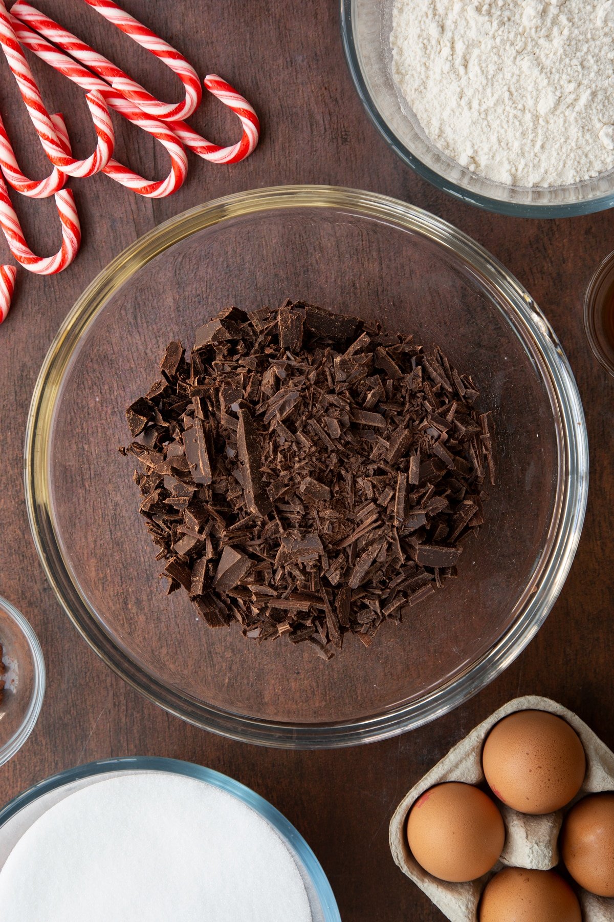 Chopped dark chocolate in a glass bowl. Ingredients to make Candy cane brownies surround the bowl.