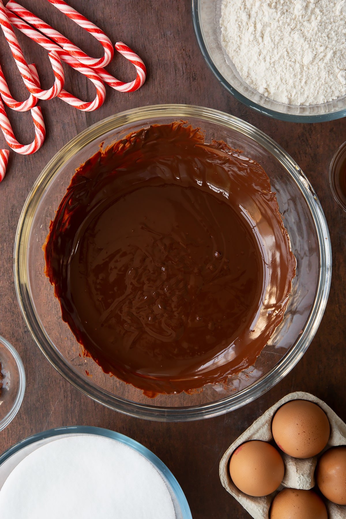 Melted dark chocolate in a glass bowl. Ingredients to make Candy cane brownies surround the bowl.