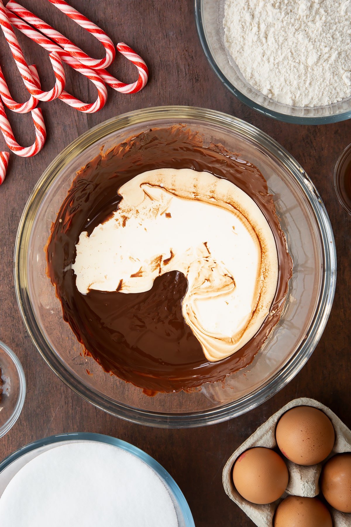 Melted dark chocolate and cream in a glass bowl. Ingredients to make Candy cane brownies surround the bowl.