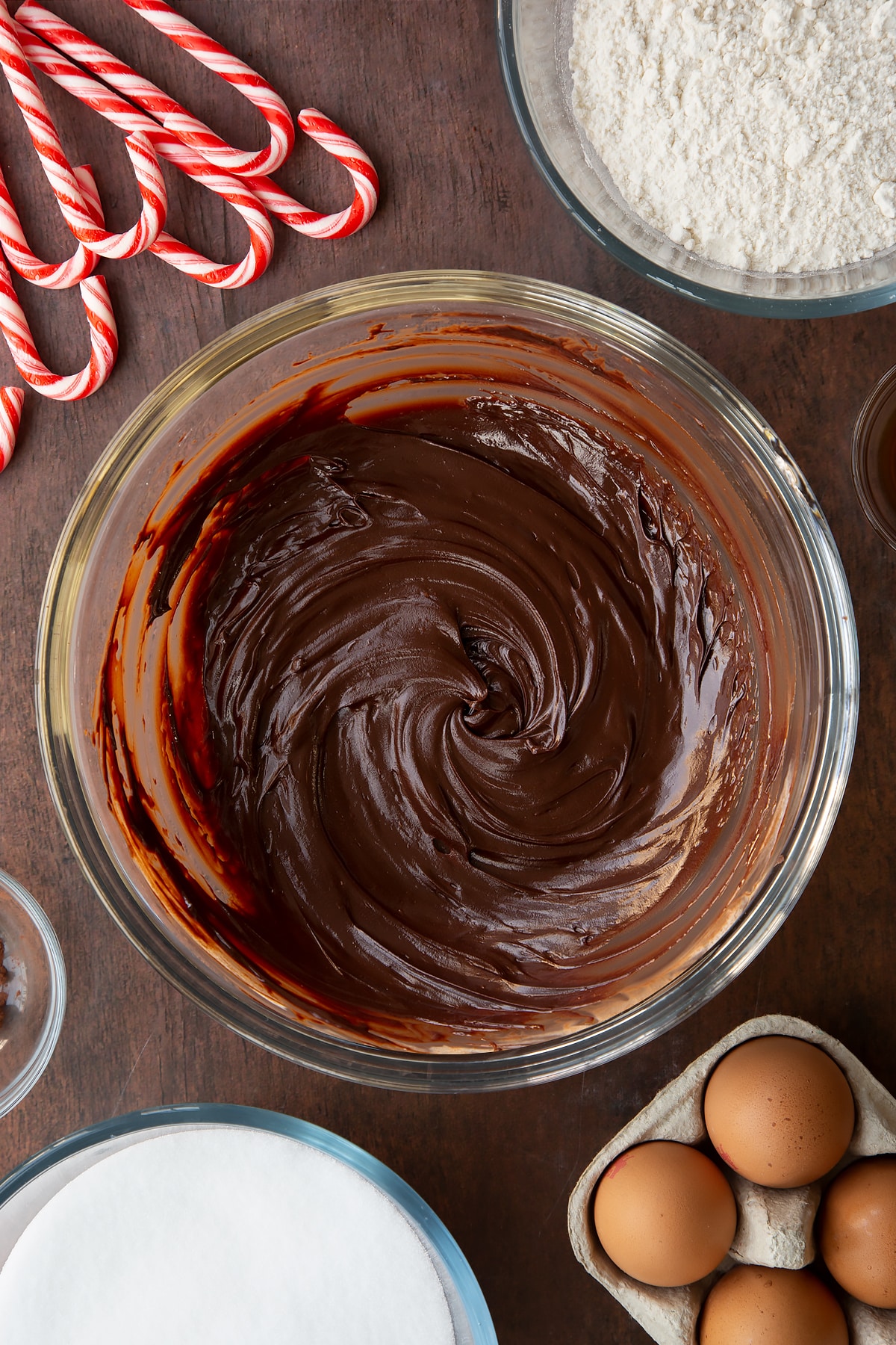 Dark chocolate ganache in a glass bowl. Ingredients to make Candy cane brownies surround the bowl.