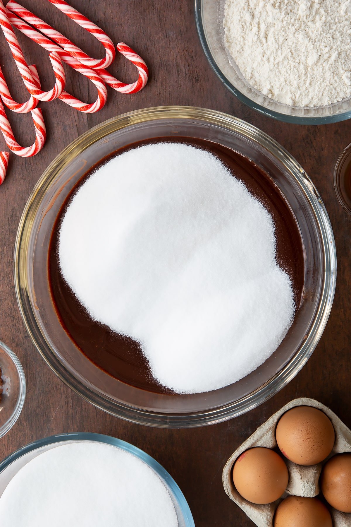 Melted butter and dark chocolate in a glass bowl with sugar on top. Ingredients to make Candy cane brownies surround the bowl.