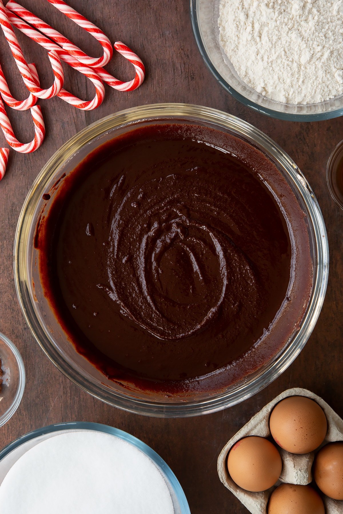 Melted butter, dark chocolate and sugar in a glass bowl. Ingredients to make Candy cane brownies surround the bowl.