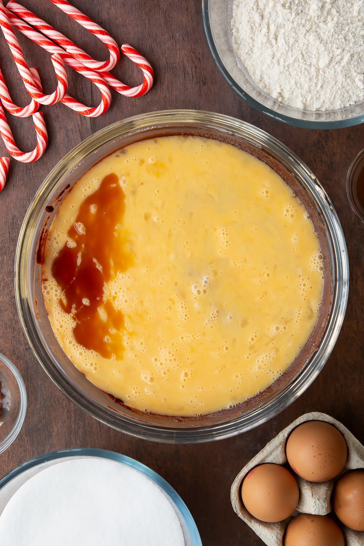 Melted butter, dark chocolate and sugar in a glass bowl with beaten eggs and vanilla on top. Ingredients to make Candy cane brownies surround the bowl.