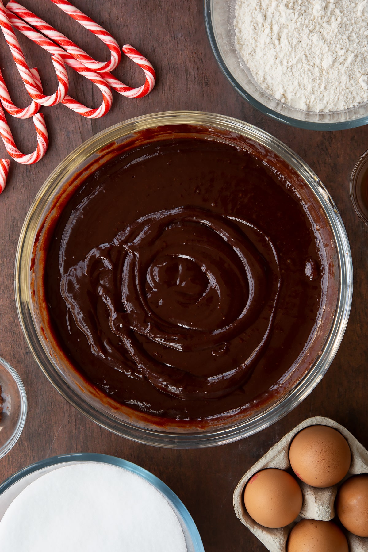 Melted butter, dark chocolate, sugar, eggs and vanilla in a glass bowl. Ingredients to make Candy cane brownies surround the bowl.