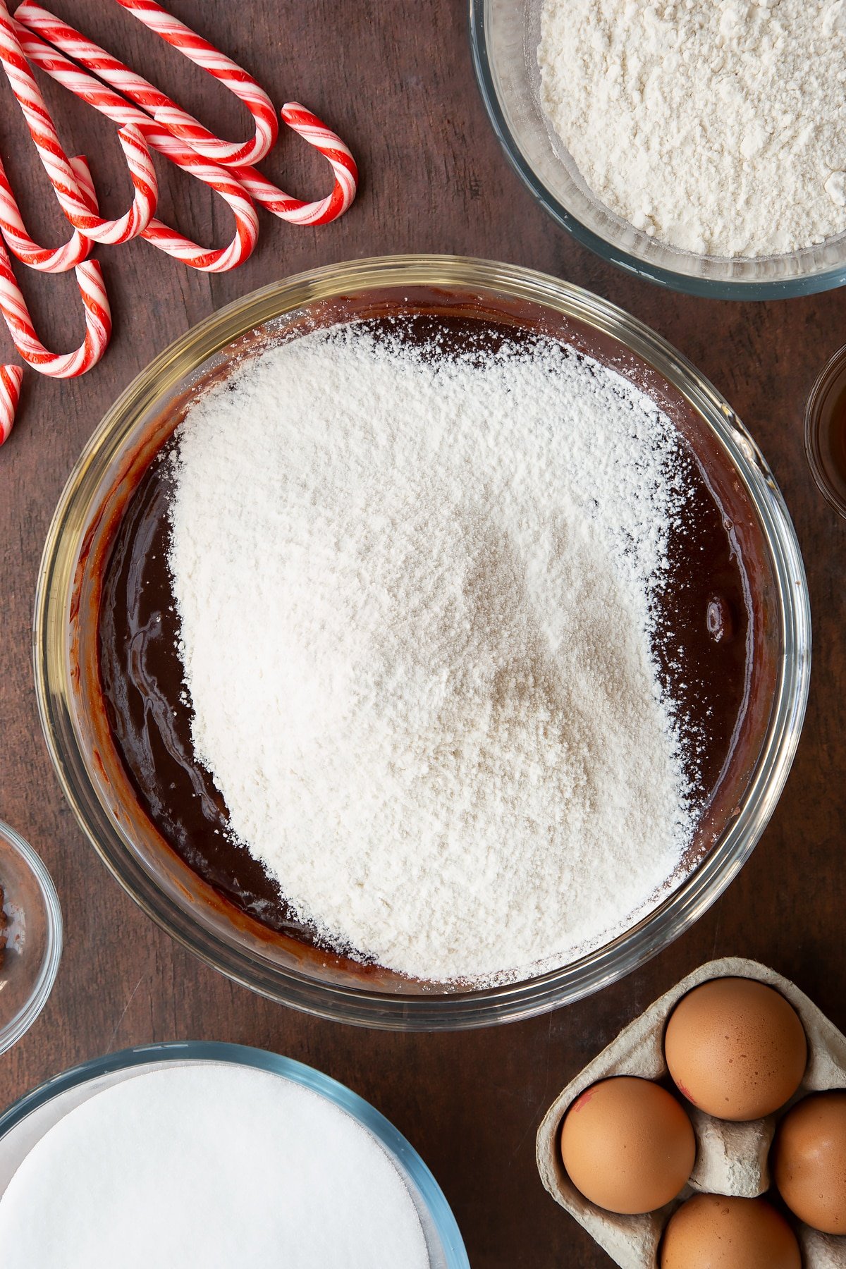 Melted butter, dark chocolate, sugar, eggs and vanilla in a glass bowl with flour on top. Ingredients to make Candy cane brownies surround the bowl.