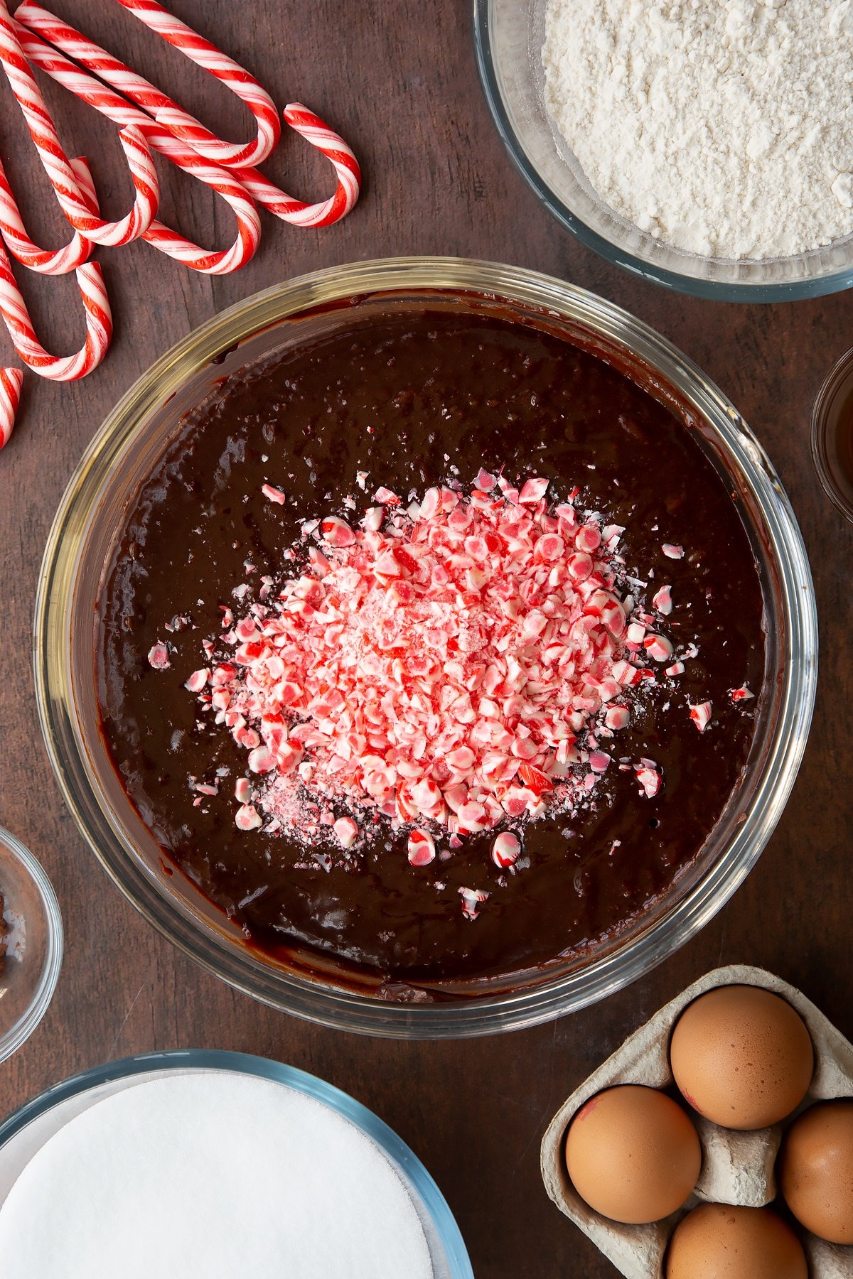 Brownie batter in a glass bowl with chopped candy canes on top. Ingredients to make Candy cane brownies surround the bowl.