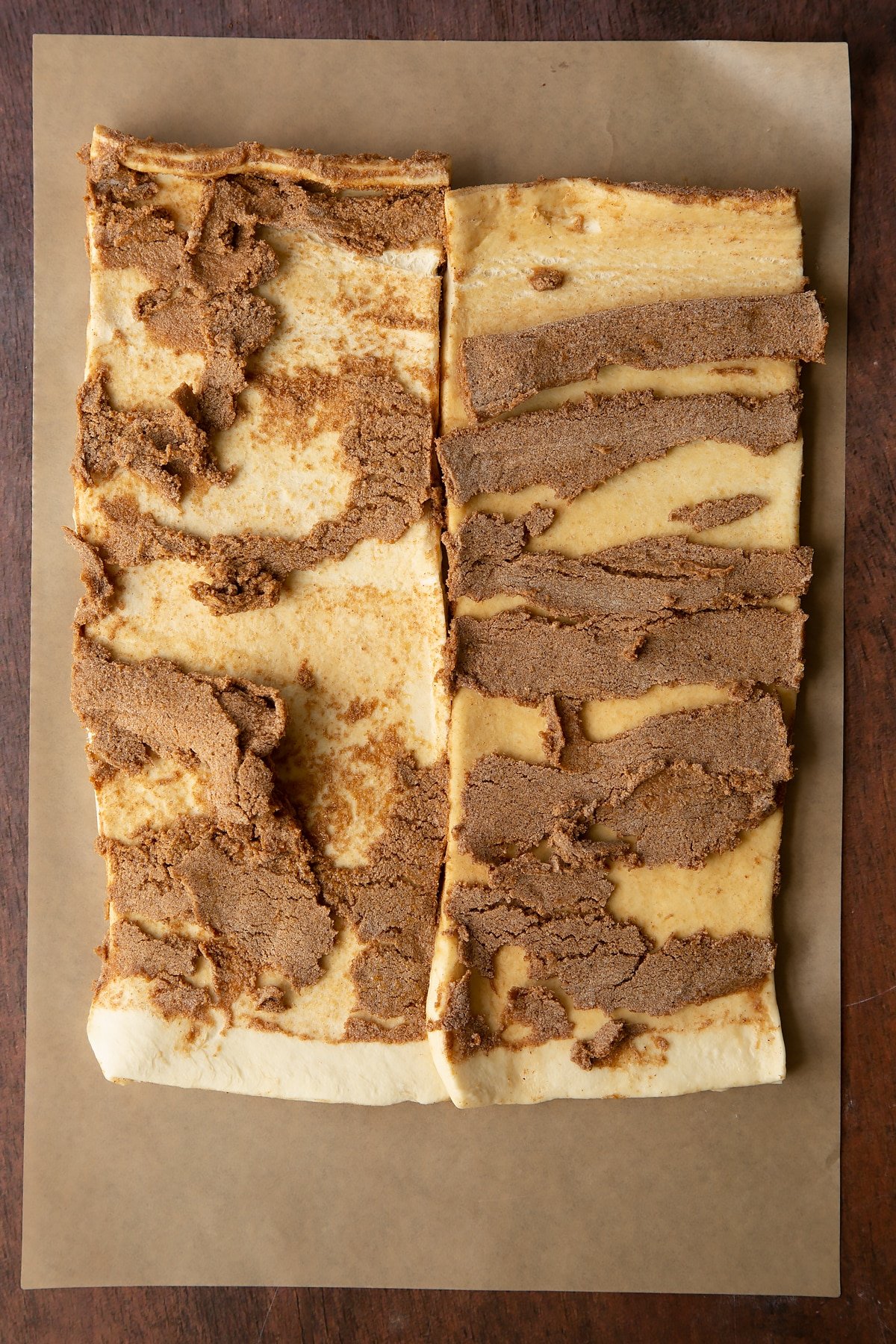 Two strips of unrolled cinnamon swirl dough on a piece of brown baking paper. The cinnamon butter filling is unevenly distributed.